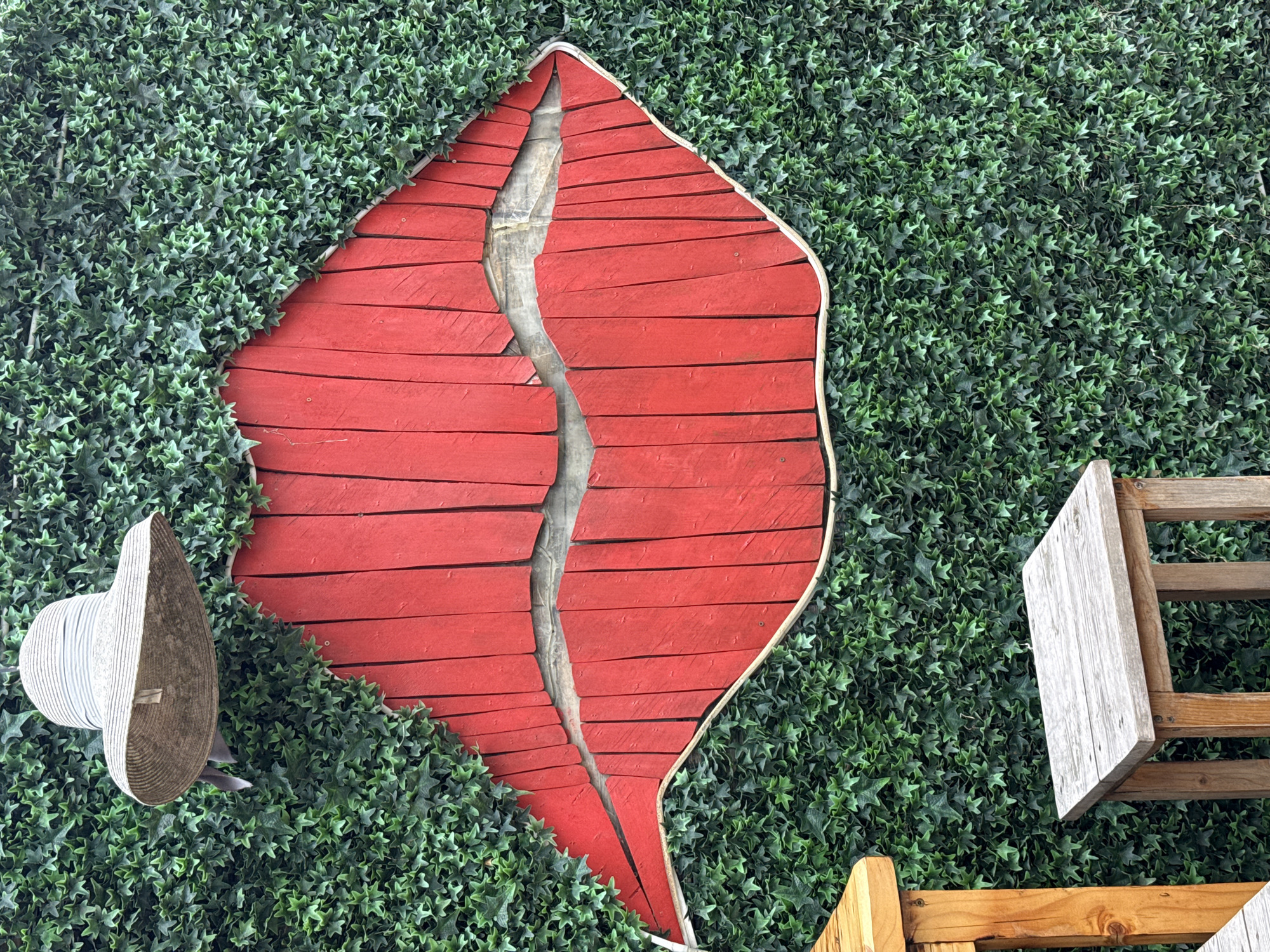 The image shows a large red wooden sculpture of lips embedded in a green ivy-covered wall. A light-colored straw hat with a dark band hangs from the top left of the wall. In the foreground, two wooden stools are partially visible, one to the left and one in the center. The scene appears to be an outdoor setting. The presence of the ivy-covered wall and the stools suggests this could be a decorative element at an outdoor cafe or a public art installation. The style of the hat and the vibrant red of the lips suggest a playful or artistic theme. This installation is located in Burgas, Bulgaria.