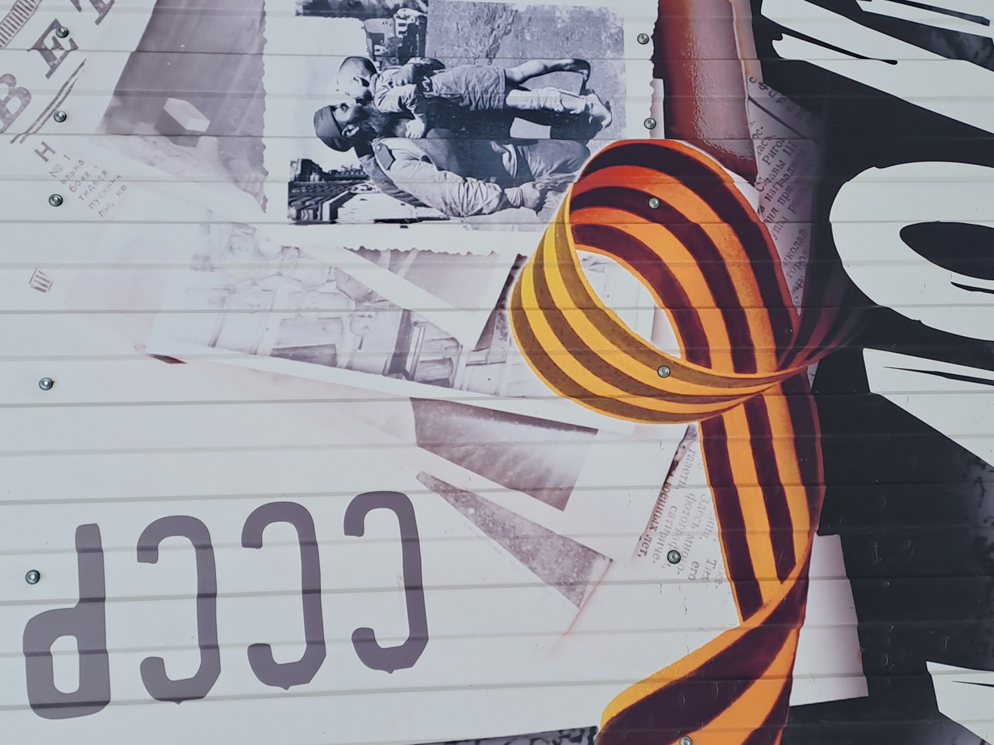 The image depicts a section of a mural or painted wall featuring historical Russian and Soviet imagery. Visible elements include:
1. **Historical Photograph:** A black and white photo shows a soldier holding a child, seemingly in a wartime or post-conflict setting.
2. **St. George Ribbon:** A distinctive ribbon with orange and black stripes, a symbol associated with Russian military valor, particularly in the context of World War II and other conflicts.
3. **"CCCP" Text:** Large Cyrillic letters "CCCP" (USSR in Russian) are prominently displayed, referring to the former Soviet Union.
4. **Text Snippets:** Fragments of Russian text from what appears to be newsprint or documents are visible.
5. **Geometric Shapes:** Simplified representations of aircraft wings or similar shapes.
6. **Overall Composition:** The image is a montage of these elements, giving a layered, collage-like feel.
Given the presence of "CCCP" and the St. George Ribbon, the mural likely commemorates Soviet military history, especially World War II.