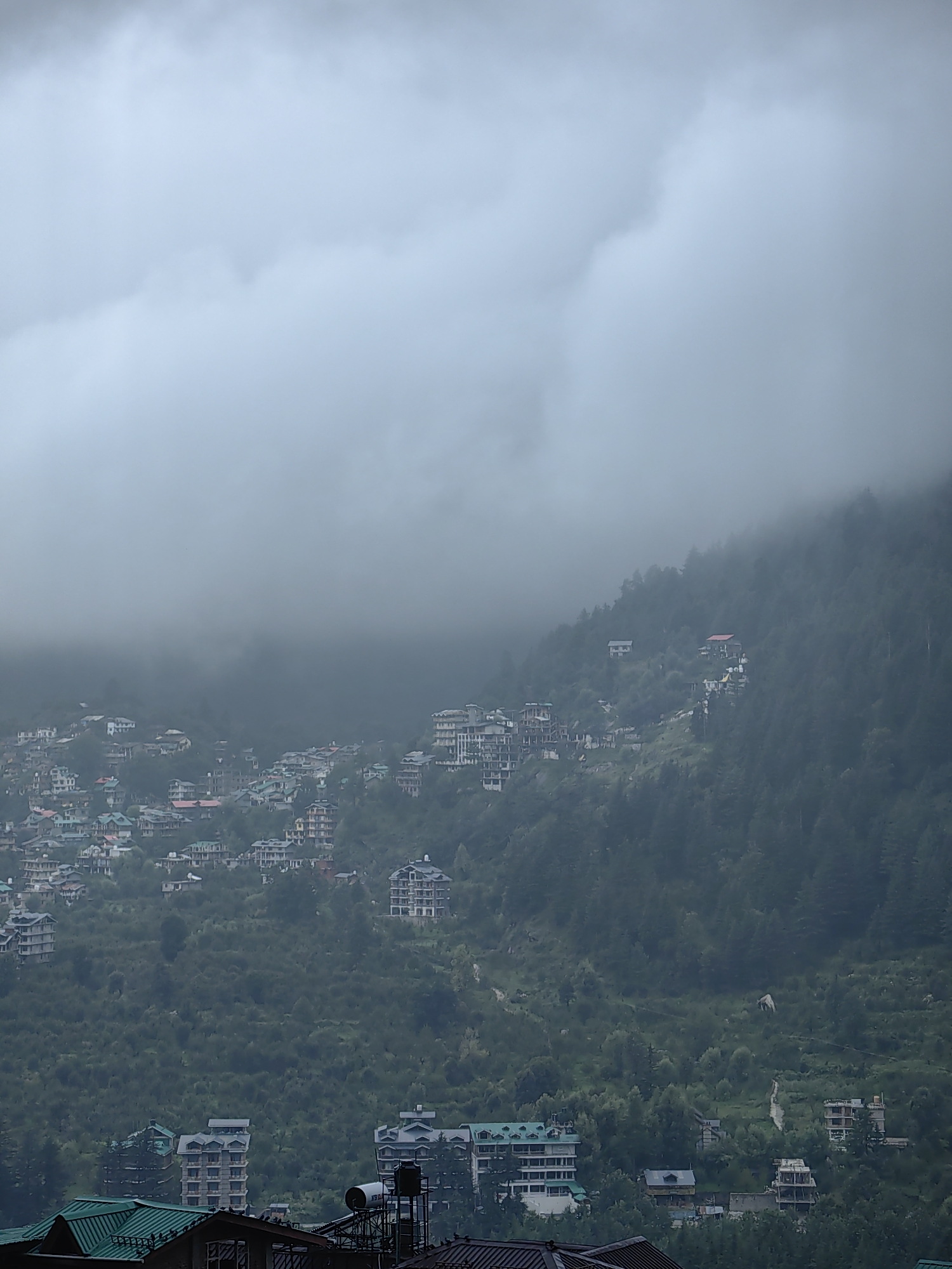 The image shows a misty view of Vashisht, India, a town in the Kullu district of Himachal Pradesh, known for its hot springs. Buildings, many of them multi-story hotels and residential structures, are nestled into the steep, forested hillside. The architecture appears to be a mix of modern and traditional styles, with many buildings featuring flat roofs and balconies. The landscape is dominated by lush green trees, which are partially obscured by a thick blanket of fog and clouds, creating a dramatic and ethereal atmosphere. In the lower part of the frame, more buildings are visible, some with green-tiled roofs. The overall impression is one of a remote mountain town shrouded in mist, with a dense, green environment.