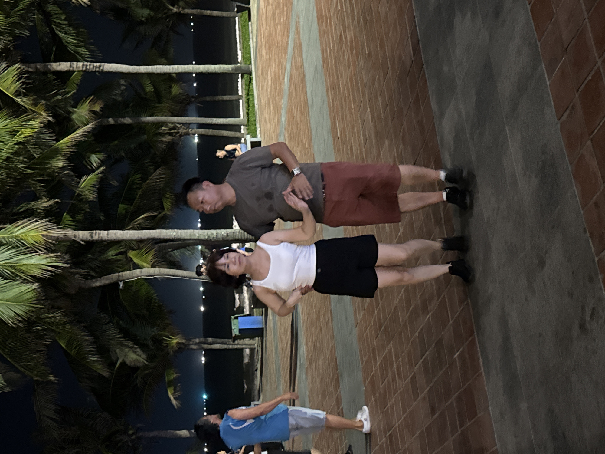The image shows a couple standing together on a tiled walkway with palm trees lining the background, suggestive of a tropical coastal area. The man is wearing a grey shirt and red shorts, while the woman is wearing a white tank top and black shorts. Another person is visible further down the walkway wearing a blue shirt. The setting appears to be at night, with lights visible in the distance, potentially indicating a seaside promenade in Đà Nẵng, Vietnam.