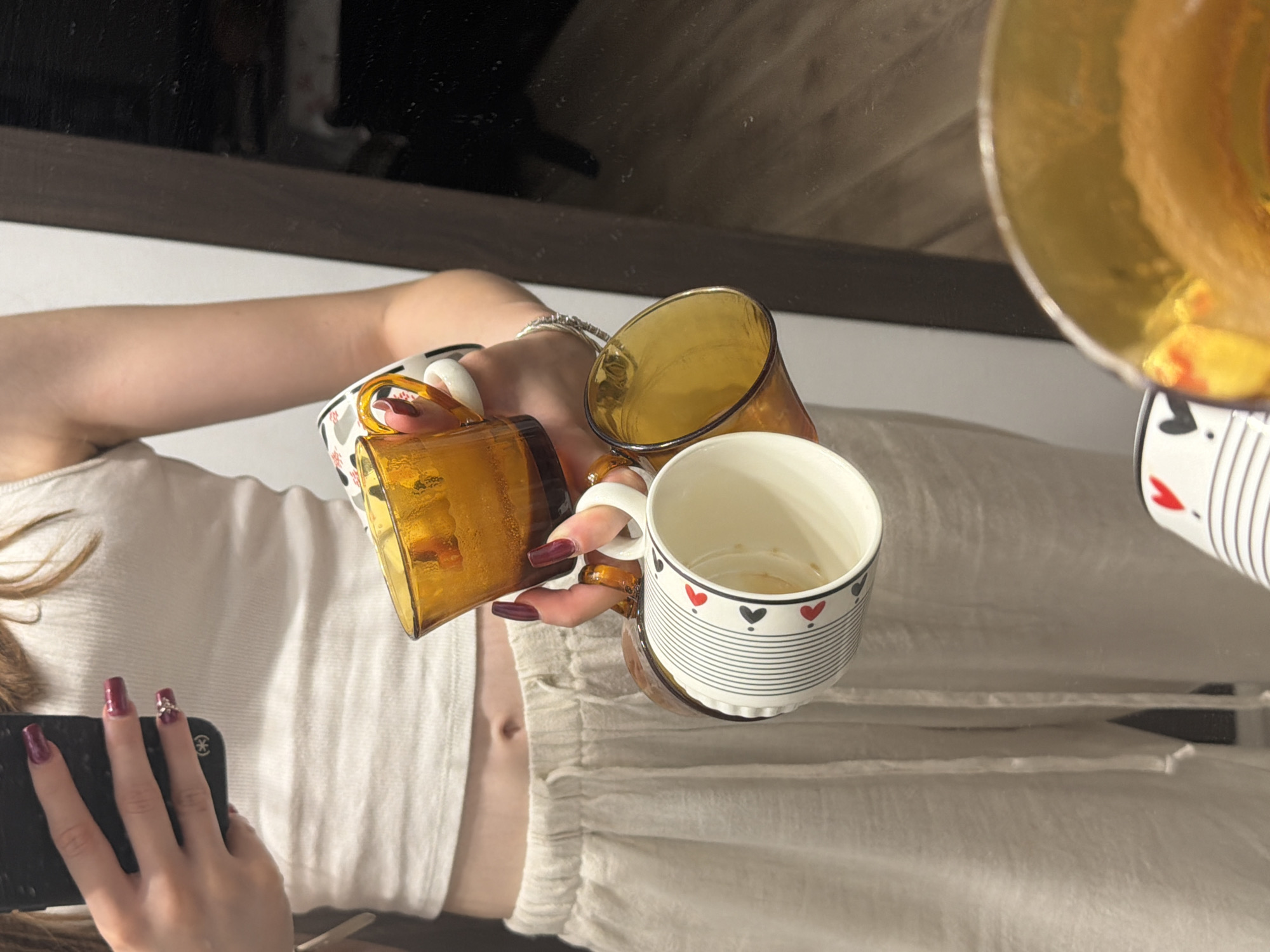 A person is holding three mugs in front of a mirror. The person is wearing a white tank top and light-colored drawstring pants. They have dark red nail polish. The mugs are amber-colored glass and white ceramic with a heart pattern. The image appears to be taken indoors, possibly in a home. There is no indication of a specific location in Varna, Bulgaria.