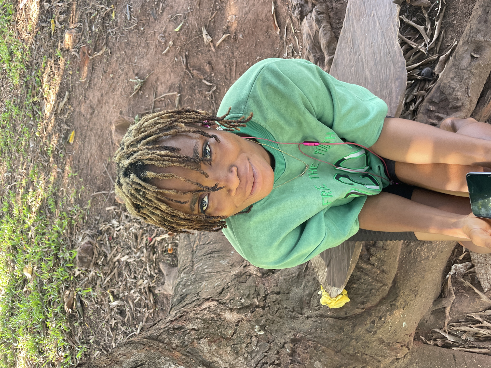 A medium eye-level shot depicts a person with light-brown dreadlocks wearing a light-green sweatshirt, sitting on a tree trunk with exposed roots. The person is wearing pink headphones and a gold necklace, and holding a smartphone in their right hand. The tree trunk occupies the lower half of the image. The ground adjacent to the tree is covered in dried leaves and sparse green grass, typical of the environment in Ogbe, Nigeria. The location appears to be a natural area possibly within the city limits of Ogbe, Nigeria.