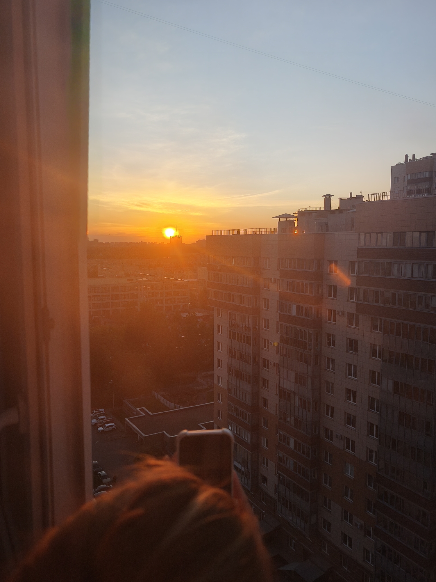 The image depicts a sunset over a city, likely taken from an elevated position such as an apartment window. The sun is visible on the horizon, casting a warm, orange light over the cityscape. The sky transitions from blue to orange. In the foreground, part of a window frame and the back of a person's head are visible. The person is holding a mobile phone, presumably taking a picture of the sunset. In the middle ground, there is a residential building complex with a grid-like pattern of windows. Below there are several parked cars outside of a building. The location is in the city of Unknown, Unknown.