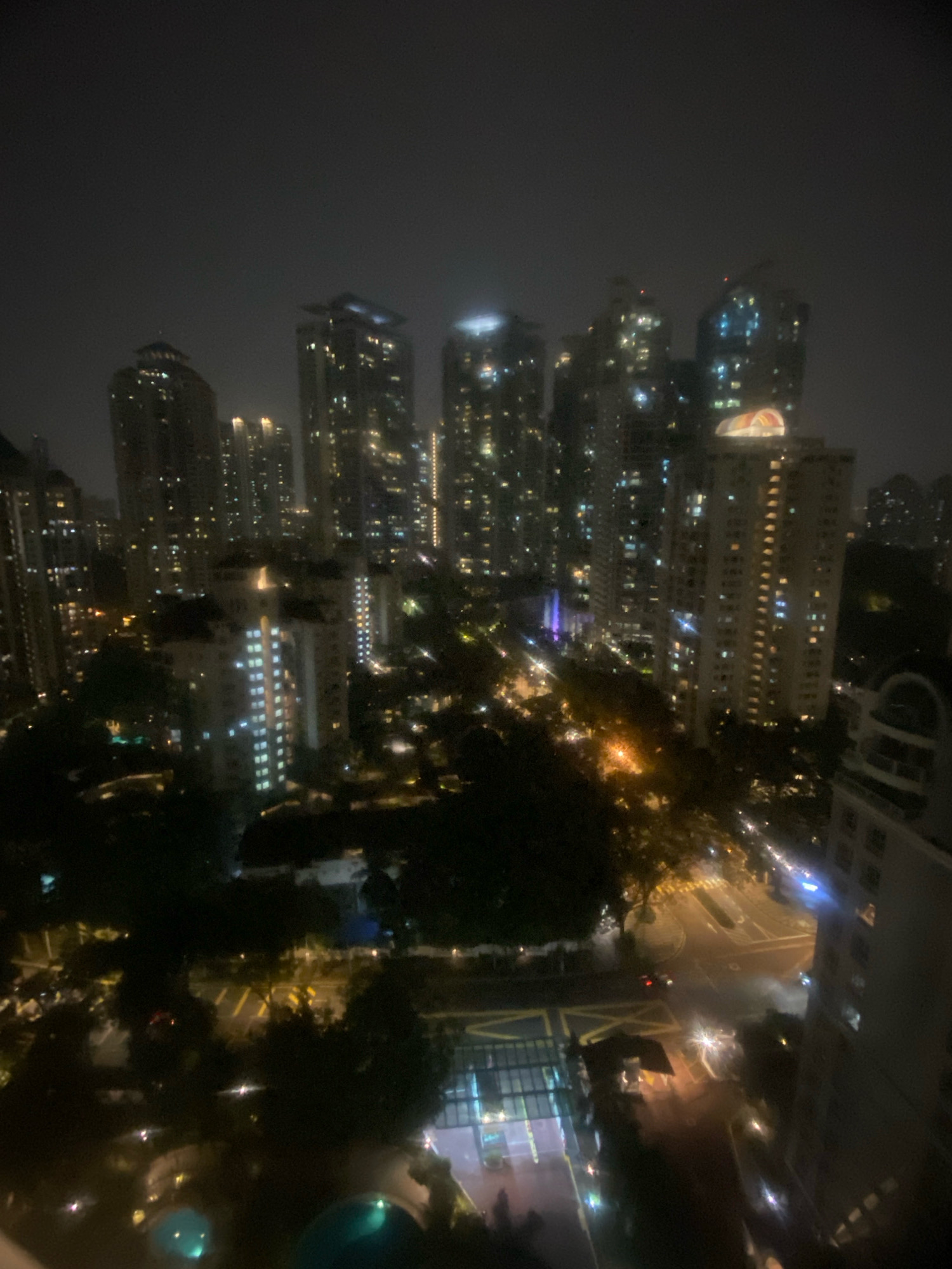 An aerial, night-time view of a city skyline in Kuala Lumpur, Malaysia. The scene is dominated by numerous tall buildings, illuminated with various lights, indicating a dense urban environment. Trees and some darker, lower structures are visible between the skyscrapers, suggesting a mix of development with green spaces. Streets with car lights are present, and some buildings have distinct architectural features, such as glowing roofs and patterned windows. The overall atmosphere is of a vibrant, well-lit city at night, possibly experiencing some haze or fog.