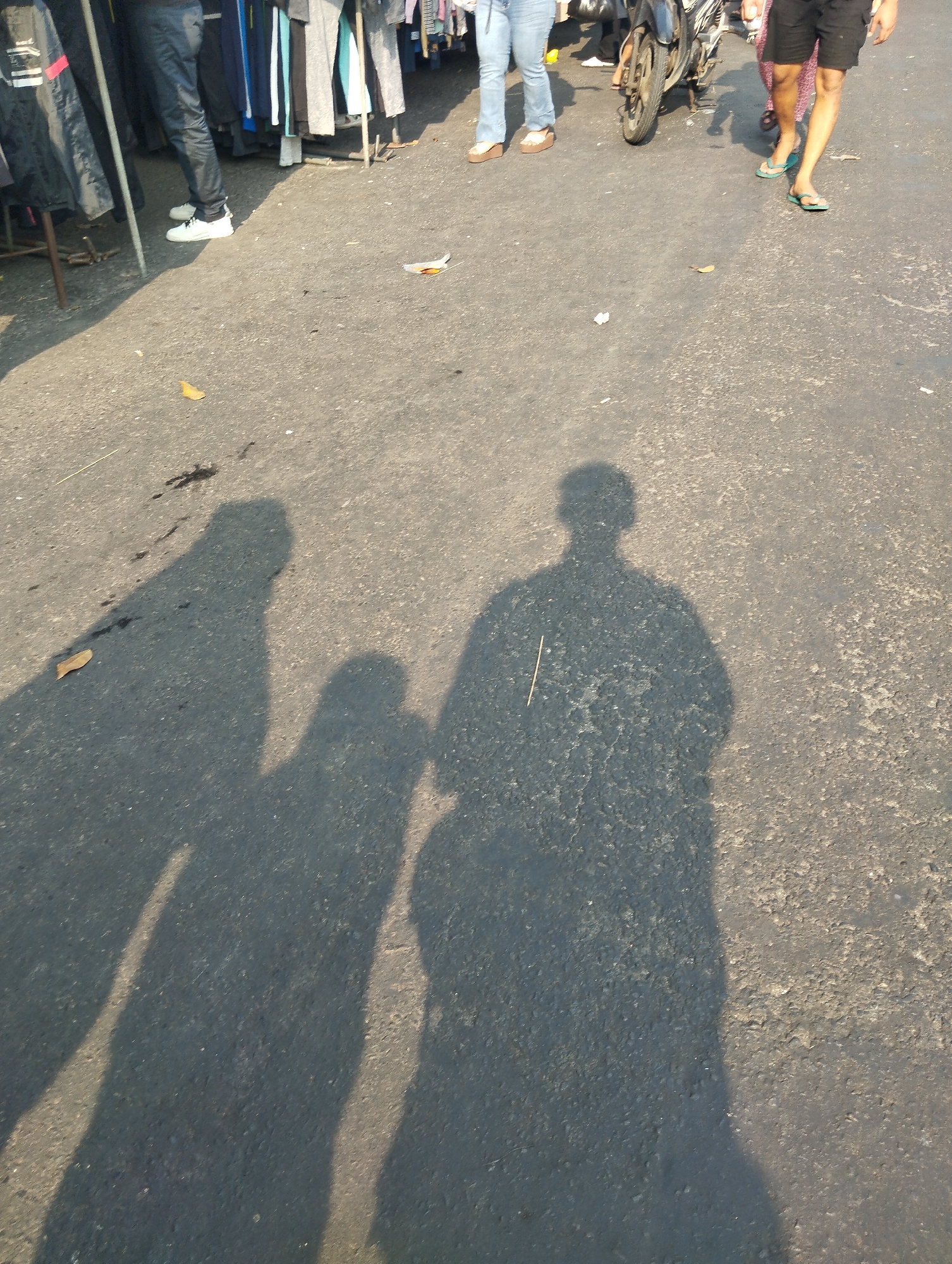 In an outdoor setting, possibly a street market in Surabaya, Indonesia, a person is captured from the knees down. They are wearing dark pants and white sneakers, standing in front of a rack of clothes. The ground is asphalt, with shadows from people and clothing displays cast across it. Further in the background, other individuals can be seen walking, one with a motorcycle. The sunlight suggests it is daytime.