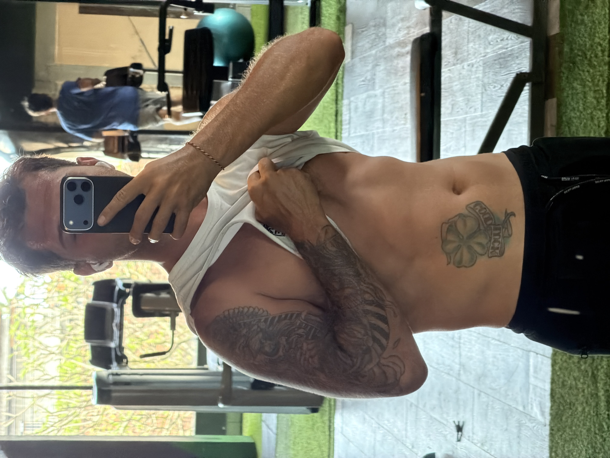 A male is in a gym in Peliatan, Indonesia, lifting his white T-shirt to reveal his torso. He has a four-leaf clover tattoo with the word "LUCK" written on it. He is holding a black cell phone to take a photo of himself. He also has a full arm tattoo on his left arm. In the background is a piece of exercise equipment, a mirror, and another person is partially visible in the mirror.