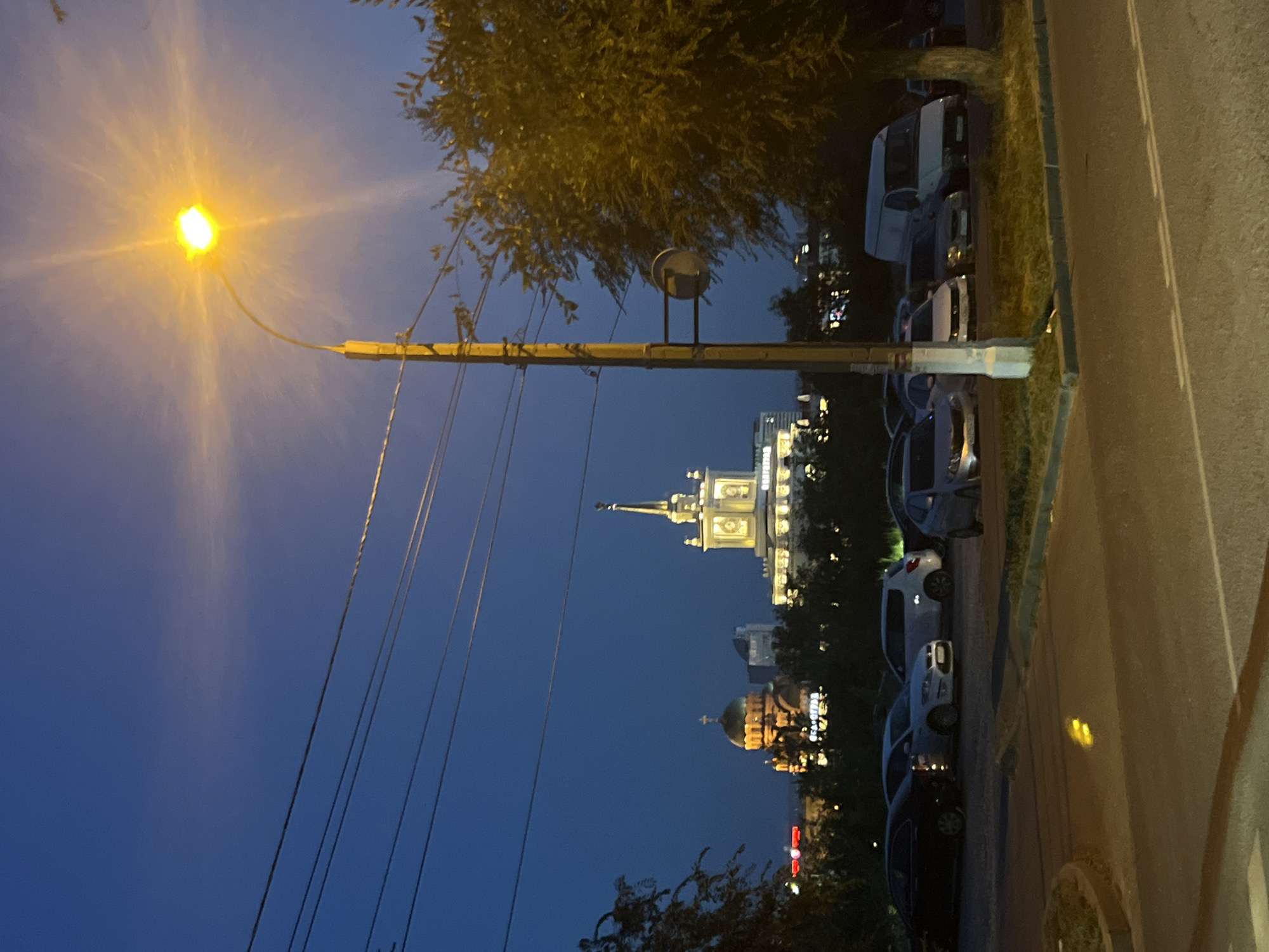 The image depicts a nighttime scene in Volgograd, Russia, featuring a parking lot in the foreground and prominent city landmarks in the background. A street light illuminates the sky with a bright yellow glow. Several vehicles are parked in the lot, including a white van, a black sedan, and other cars. In the middle ground, a large building with a spire and illuminated windows stands out, appearing to be a landmark or a significant structure. To the left, a golden-domed building, likely a church, is visible. The sky is a deep blue, suggesting dusk or early night. Power lines are strung across the upper portion of the frame. Trees are present on the right side of the image, their foliage illuminated by the street light. The overall scene conveys an urban setting at night, with a mix of modern and traditional architecture.