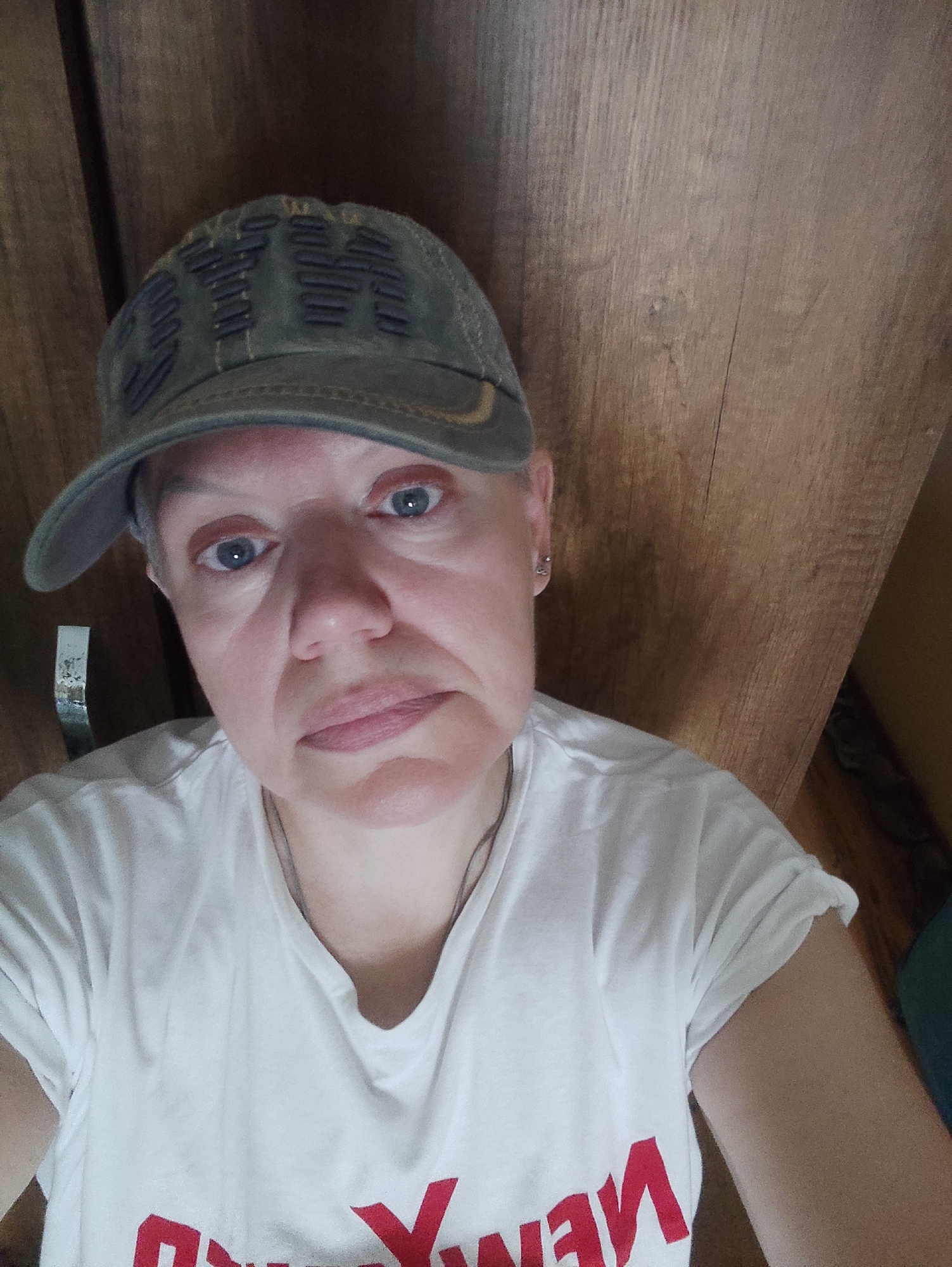 A woman wearing a green baseball cap and a white t-shirt with red lettering is looking at the camera. She has short, grey hair and blue eyes. The background is a wooden wall. The image appears to be a selfie taken indoors. There is no indication of the location being Kadıköy, Turkey.