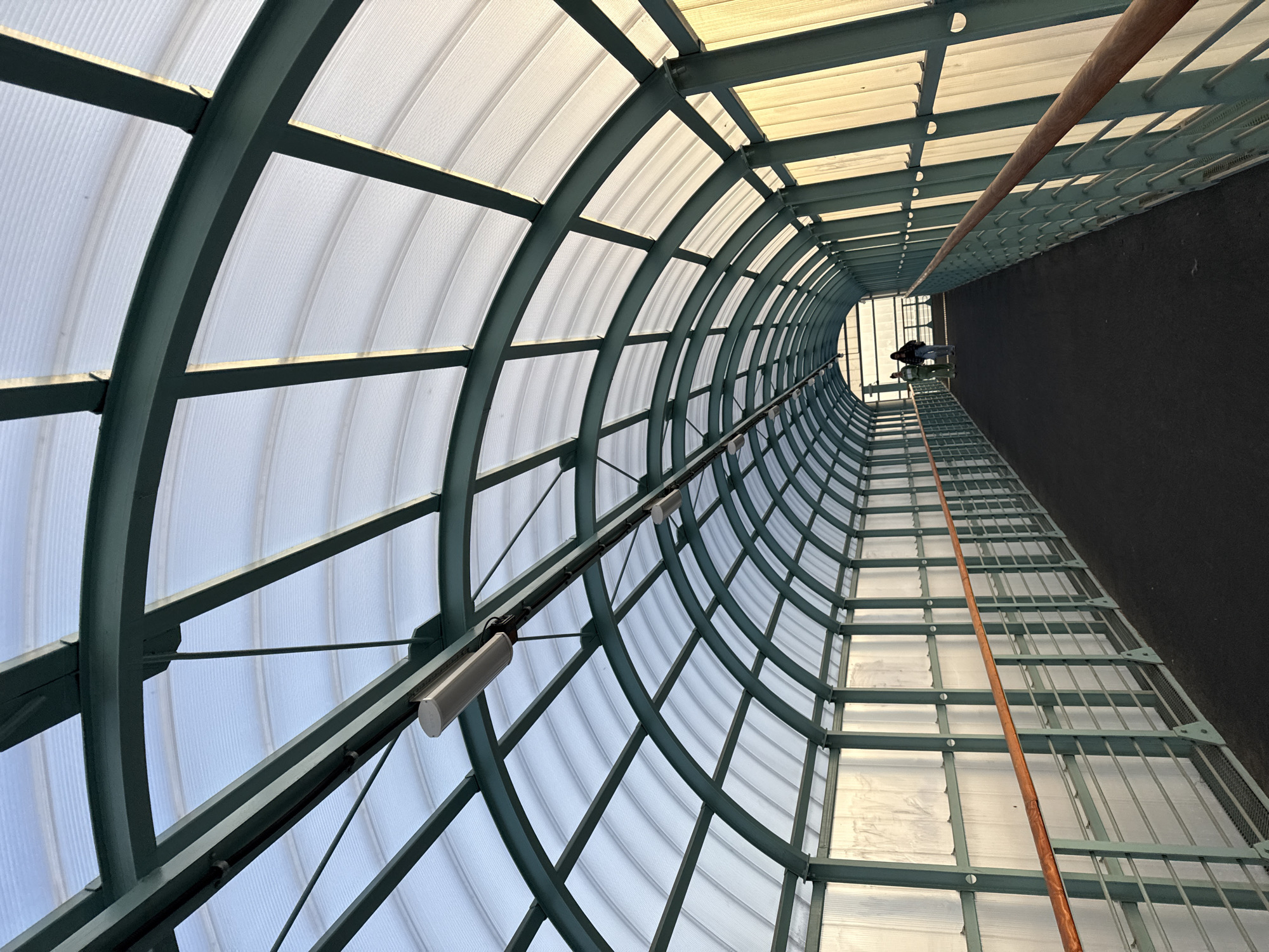 The media file depicts the interior of a covered walkway or tunnel. The structure is characterized by a curved, arched roof made of translucent, ribbed panels supported by a green metal framework. The walkway has a dark, possibly asphalt, floor on one side, and a metal grate walkway on the other, suggesting a multi-level or complex structure. A wooden handrail runs along the length of the walkway.
There is one person visible in the distance, walking away from the camera on the grated walkway. They are wearing dark clothing. Two cylindrical light fixtures are attached to the metal structure of the roof, positioned along the length of the walkway.
The lighting suggests it is daytime, with natural light filtering through the translucent roof, creating a diffused glow. There are no obvious weather cues, but the enclosed nature of the walkway implies protection from the elements. The perspective is a low-angle shot looking upwards and down the length of the tunnel, emphasizing the architectural design and the sense of depth. The overall mood is one of urban passage or infrastructure, with no strong emotional cues evident. There is no visible text in the image. The location context provided is Perm, Russia.