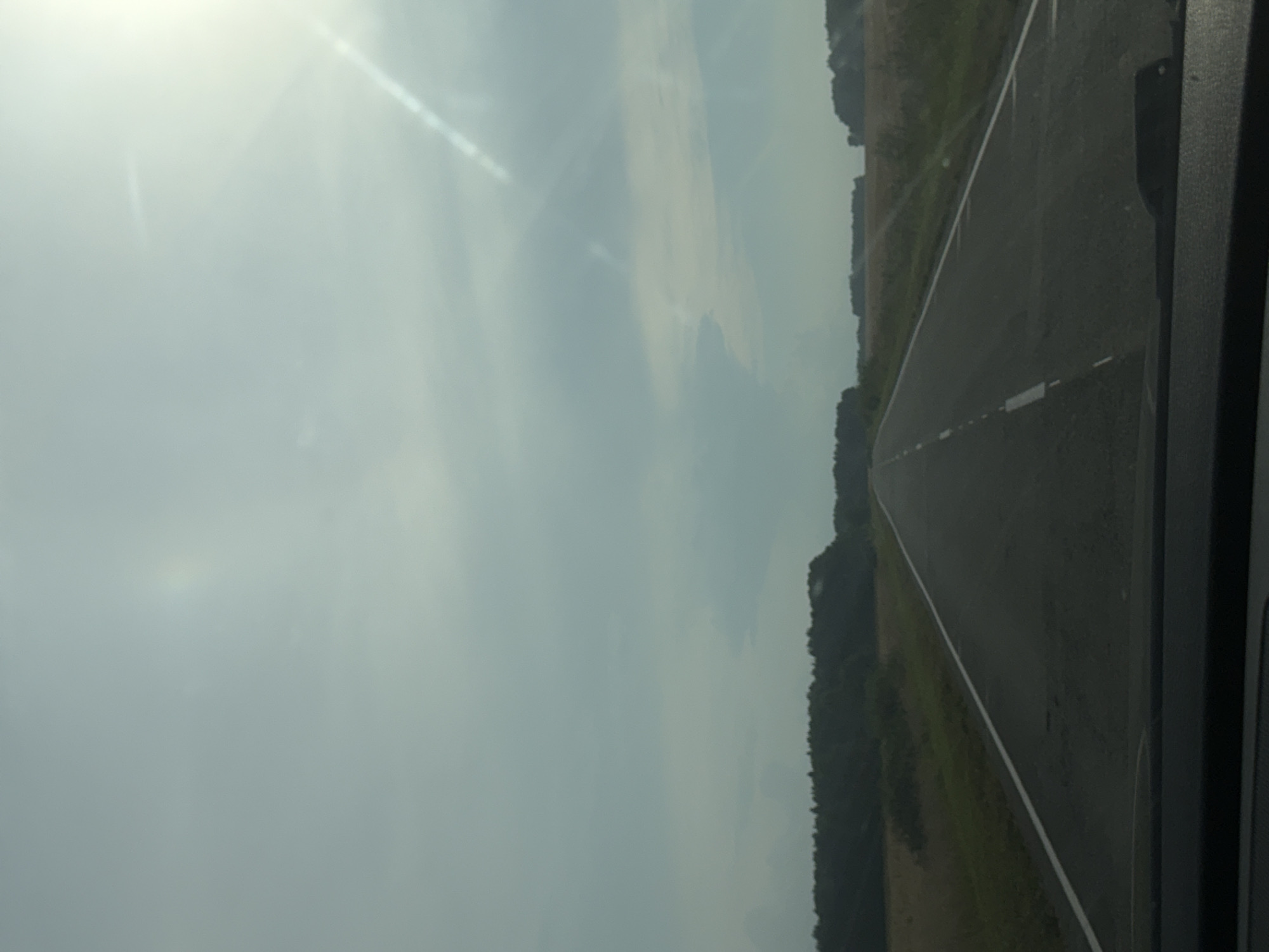The image depicts a straight, paved road with a white dashed line marking the center. The road stretches into the distance, bordered by grassy verges and fields on either side. In the background, a dense line of dark green trees forms a horizon, with more distant, lighter green foliage visible. The sky above is overcast with a pale blue hue, and bright sunlight streams in from the upper right corner, creating lens flares. The perspective is from inside a vehicle, with the dashboard visible in the foreground. There are no people or animals present in the scene. This scene is a rural landscape on a road in Nylga, Russia.