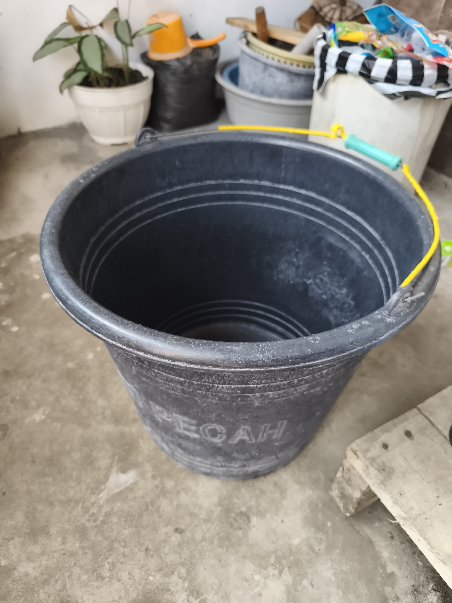 A black plastic bucket with a yellow rope handle lies on a concrete floor in what appears to be an outdoor or semi-outdoor area. The bucket has horizontal ridges around its exterior and some white residue on its surface. The word "PECAH" is embossed on the side of the bucket. In the background, a white pot containing a green plant sits on the left. Behind the bucket, several other containers are visible, including a black woven bag with an orange scoop on top, and a large white bin with a black and white striped cloth draped over it. To the right of the bucket, a wooden plank is propped up. The lighting suggests it is daytime. The location is described as a city in Yogyakarta, Indonesia.