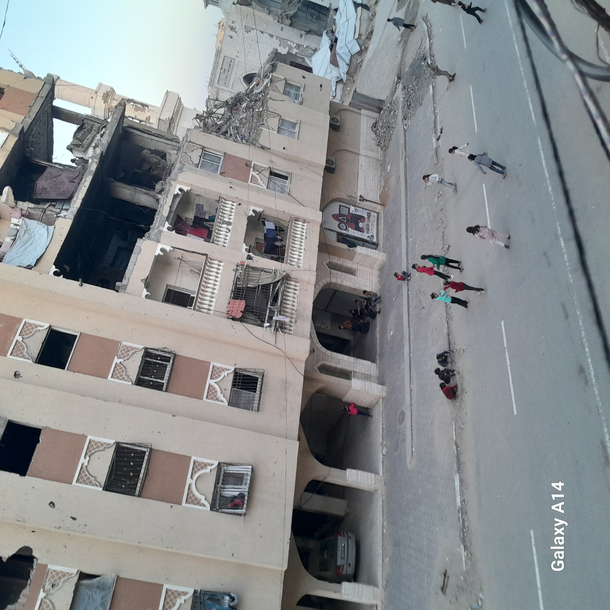 The image shows a partially destroyed building in an urban setting. The upper floors of the building have significant structural damage. Debris litters the ground near the building and the adjacent street. There are several people on the street, some standing and some lying down. There is a damaged car parked in a shaded area of the building. The location is in the city of Unknown, Unknown.