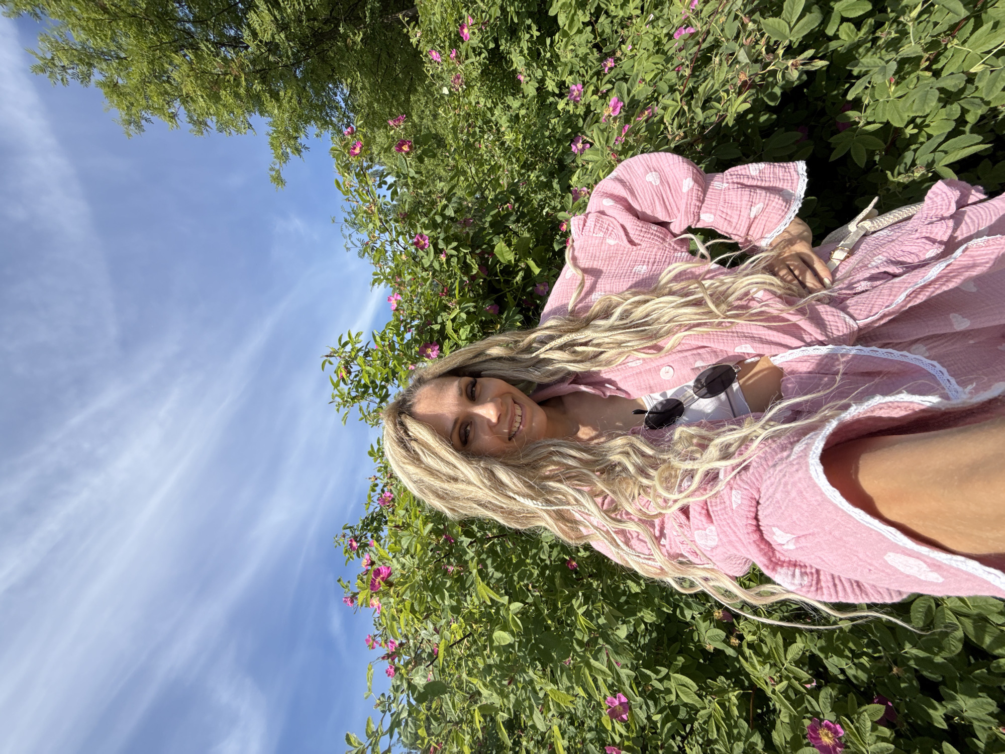 An adult female is captured in a waist-up selfie, smiling directly at the camera, in an outdoor setting within the city of Rusanovo, Russia. She possesses long, textured, light blonde hair that extends past her shoulders. Her face displays a broad smile, with visible freckles across her nose and cheeks. She wears a loose-fitting, light pink, textured button-up shirt or jacket patterned with small white hearts and featuring ruffled cuffs with white lace trim. A white crop top is visible underneath this garment. Dark-lensed sunglasses are hooked onto the collar of her pink top. Rings are visible on the fingers of her right hand, which is placed on her hip, while her left arm is extended, holding the camera in a selfie pose.
The background features dense, lush green bushes densely populated with bright pink, five-petaled flowers, indicative of wild roses. Above the vegetation, a clear blue sky is visible, traversed by wispy, white cirrus clouds. A portion of a green deciduous tree is present on the far right side of the frame, partially obscured by the flowering bushes. The scene is brightly illuminated by natural daylight.