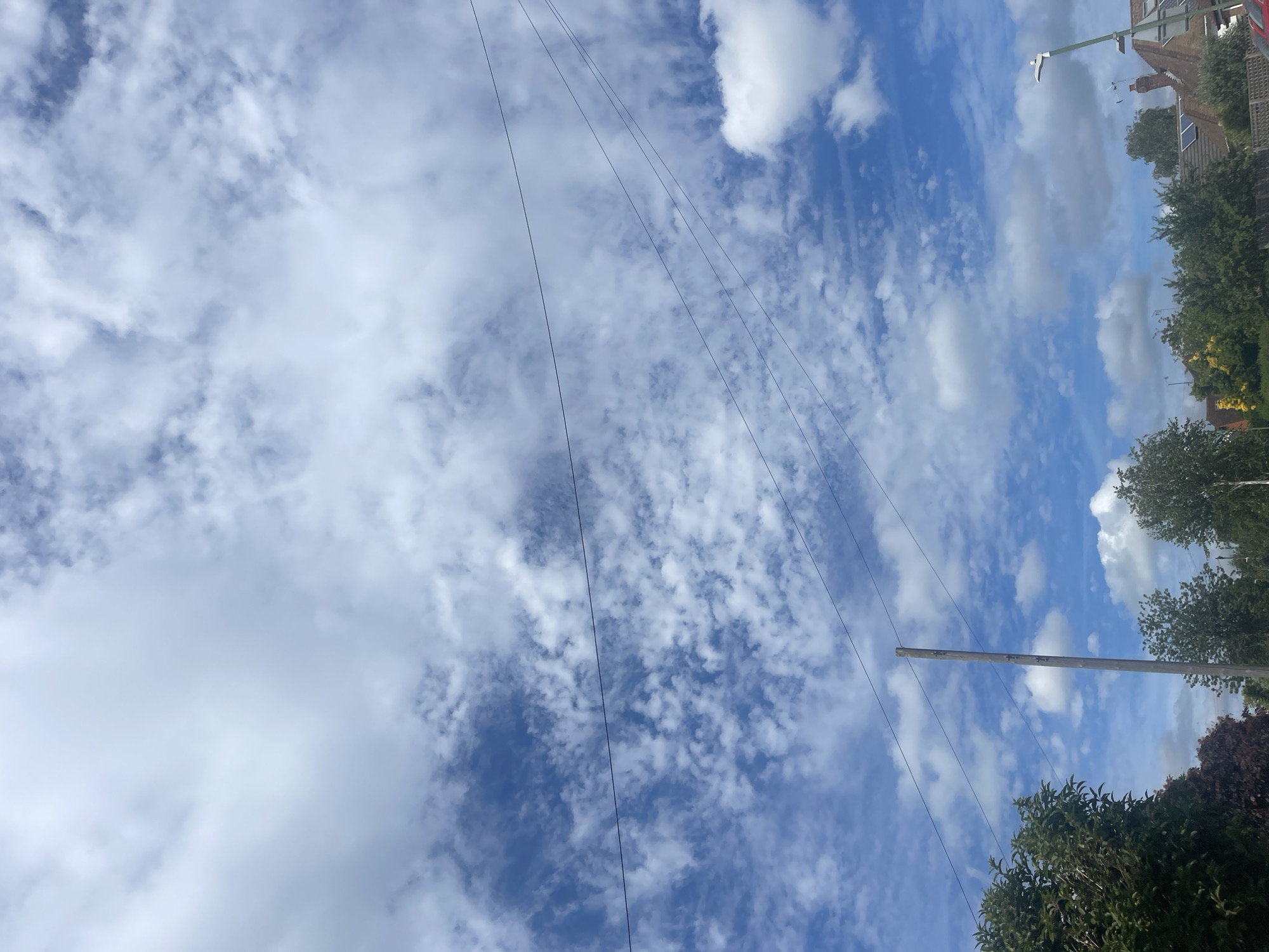 The image depicts a suburban street scene in Maidstone, United Kingdom, under a partly cloudy sky. The lower portion of the frame is dominated by trees and rooftops, indicating a residential area. A wooden utility pole stands prominently in the left-center of the image, with several wires strung between it and other unseen structures, crossing diagonally through the sky. The sky itself is a vibrant blue, with scattered white clouds of various sizes and densities, suggesting a bright day. A streetlamp is visible on the far right, further confirming the urban setting. There are no people or vehicles visible in the frame.