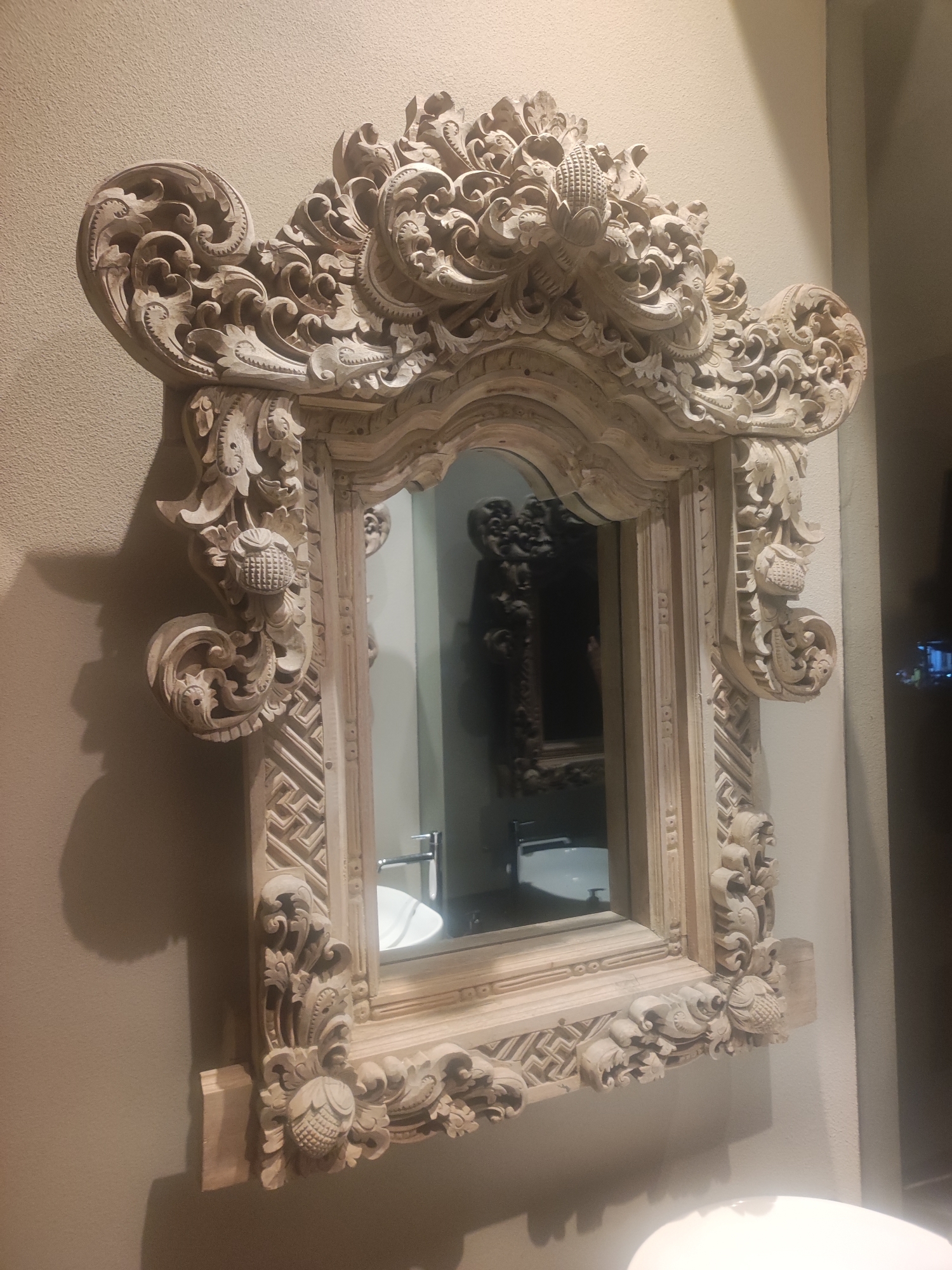 The central subject is a large, highly ornate wooden mirror with a rectangular glass panel. The mirror frame is made of light-colored wood, possibly whitewashed or natural, and is extensively hand-carved with intricate designs. These carvings feature elaborate scrollwork, stylized floral motifs, and decorative elements resembling textured pinecones or lotus buds at the corners and along the sides. The top of the frame is particularly decorative, with a central flourish and dramatic, undulating curves.
The mirror is mounted on a plain, light beige or tan wall with a subtle texture. Shadows cast by the deep carvings of the frame are visible on the wall, indicating ambient lighting.
The mirror's reflection shows parts of a modern bathroom. Clearly visible in the reflection is a white ceramic sink basin with a contemporary silver faucet. A darker, ornate object, possibly another mirror or a piece of furniture, can be faintly seen in the deeper reflection within the main mirror.
No specific activity or event is taking place; the image is a static capture of the mirror in its setting. There are no people visible. The intricate, organic carving style of the mirror frame is characteristic of traditional Balinese or Indonesian artistry, aligning with the provided location context of North Kuta, Indonesia. There is no visible text in the image.