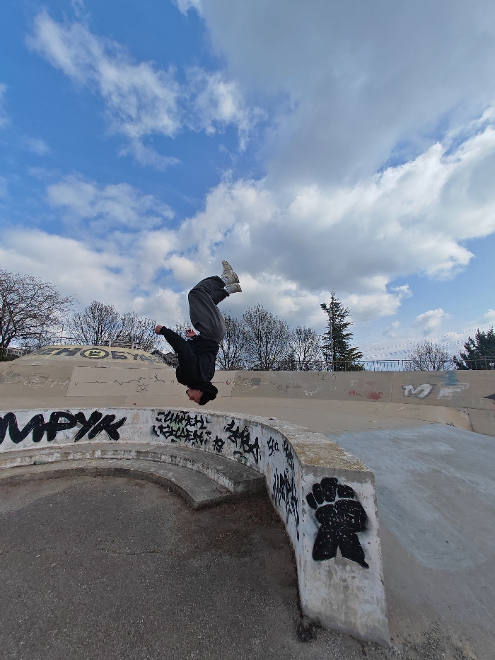 A single individual is captured in mid-air performing an inverted acrobatic flip within an outdoor skatepark in Shumen, Bulgaria. The person is dressed in a black hooded sweatshirt, dark gray loose-fitting pants, and light-colored athletic shoes with white socks, positioned head-down with legs extended upwards. The immediate foreground features a curved concrete barrier, heavily marked with black graffiti including Cyrillic script and a prominent black fist or paw print stencil. The ground is dark, paved material. In the background, a flat concrete wall is visible, also with graffiti, beyond which a line of bare deciduous trees and one evergreen tree stand against a partially cloudy blue sky.