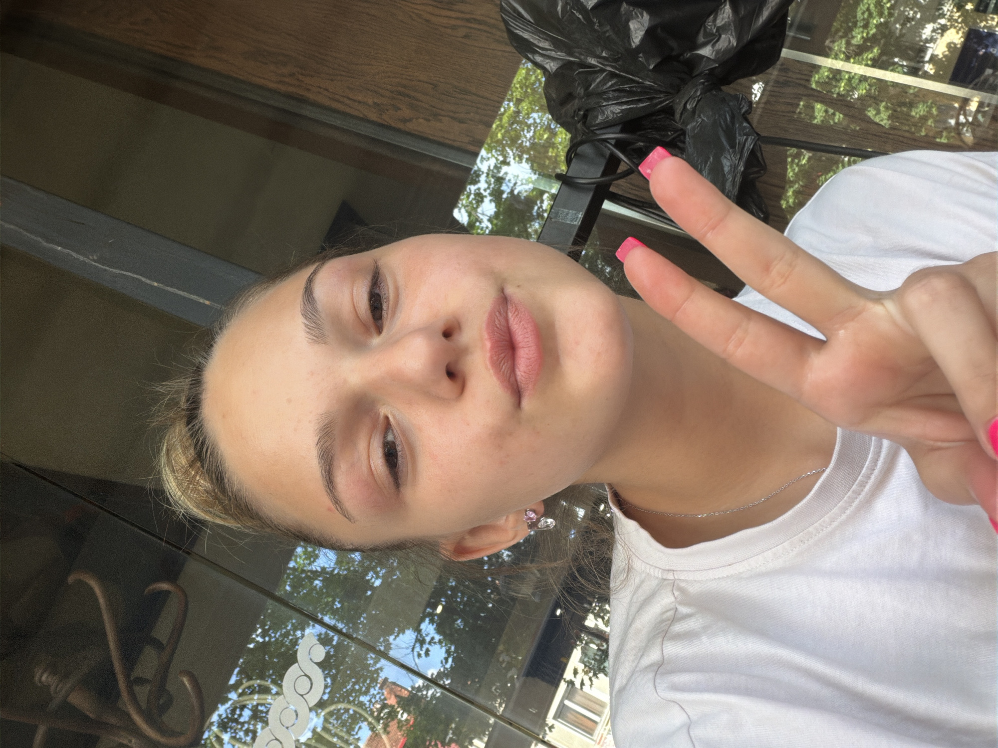A young woman poses for a selfie, making a peace sign. She has fair skin, light brown hair pulled back, and pink nail polish. She wears a white T-shirt and a thin silver necklace. Her left ear has an earring. The background includes a window reflecting trees and buildings. Outside the window, there is greenery and buildings visible. A black garbage bag is positioned behind her. The location may be a cafe or restaurant in Troyan, Bulgaria, suggested by the building and tree reflections in the window and the interior furniture visible.