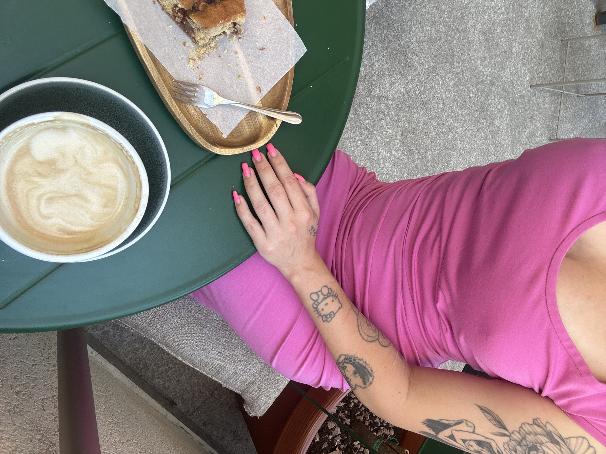 The image captures a close-up, overhead view of a person sitting at an outdoor table with a coffee and a slice of cake. The subjects are the person's arm with tattoos, a white cup of latte art coffee, a slice of cake on a wooden platter with a fork, and a pink top worn by the person. The setting appears to be a cafe or restaurant with a green table and a textured, grey ground.
The person's arm is adorned with several tattoos, including a Hello Kitty character, a hedgehog, and floral designs. Their fingernails are painted a vibrant pink. The coffee cup shows a swirl pattern in the foam, indicating it's a carefully prepared beverage. The cake appears to be a dense, possibly chocolate or nut-based confection.
The overall mood is relaxed and casual, suggesting a moment of enjoyment and leisure. The lighting implies it could be daytime, possibly mid-morning or afternoon, with natural light illuminating the scene. The background is slightly blurred, keeping the focus on the immediate items on the table and the person's arm. There is no visible text in the image. The location context of Budva, Montenegro, is provided but not directly evident from visual cues within the image itself.