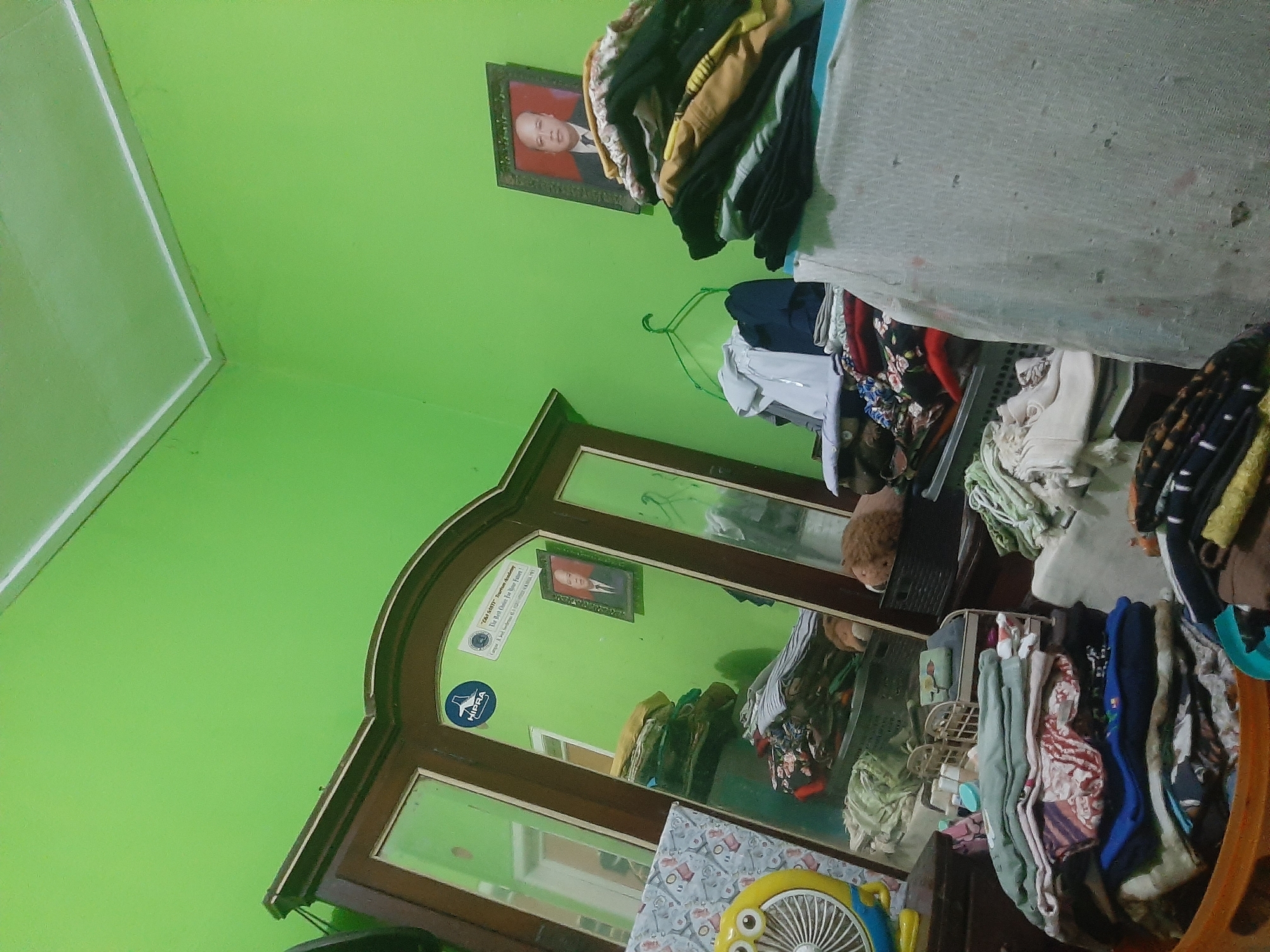 The image depicts an interior scene, likely a bedroom, with bright green walls and a white ceiling. Dominating the left side of the frame is a large, dark wooden wardrobe with a mirrored door. Reflected in the mirror are more of the green walls, an open doorway, and possibly another person, though they are indistinct. Affixed to the wardrobe door are two stickers, one circular and blue with white text and a stylized graphic, and another rectangular with text. To the right of the wardrobe, a framed portrait of a man in a suit and tie is hung on the wall. In front of the wardrobe and to the right, several stacks of folded clothing are visible, some in baskets and others on shelves or flat surfaces. A yellow fan with a cartoon face is positioned on the left side of the wardrobe. The overall impression is one of a lived-in space, possibly undergoing a tidying process or in a state of disarray due to laundry. The specific location of Sumampir, Indonesia, is not visually evident from the image itself, but the context of the request implies it is the intended setting.