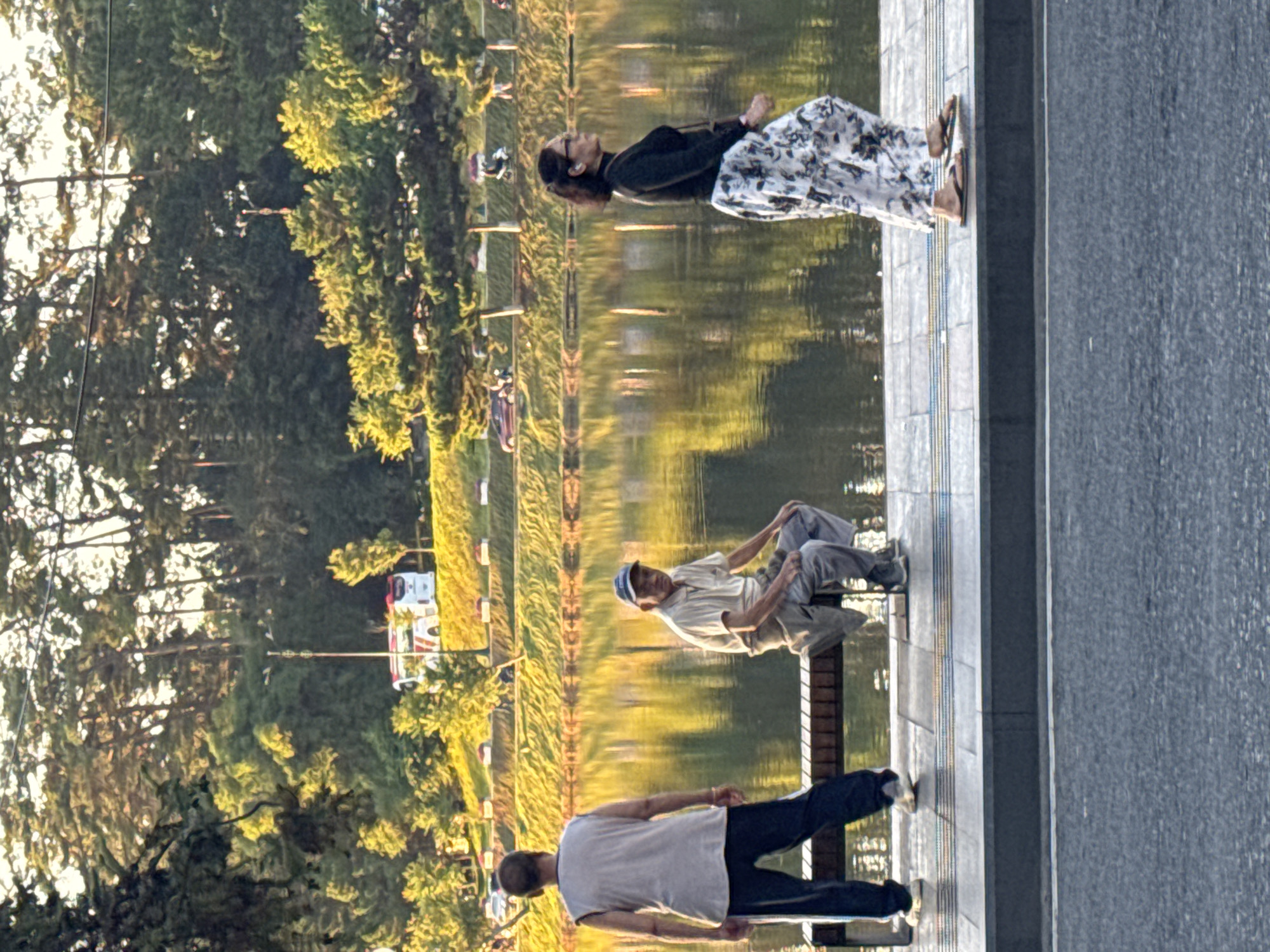 The image depicts a scenic waterside location, likely Xuan Huong Lake in Da Lat, Vietnam, bathed in the warm glow of late afternoon sunlight. In the foreground, a paved walkway runs alongside the water. Three individuals are present. On the left, a man stands with his back to the camera, wearing a light-colored sleeveless shirt and dark pants. To his right, an elderly man is seated on a bench, facing the camera, dressed in a light-colored short-sleeved shirt, grey pants, and a baseball cap. Further to the right, a woman walks away from the camera, wearing a dark long-sleeved top and a patterned skirt. Beyond the walkway and water, a tree-lined embankment slopes upward, with various vehicles including a bus visible in the distance. The water reflects the surrounding trees and sky, creating a serene atmosphere.