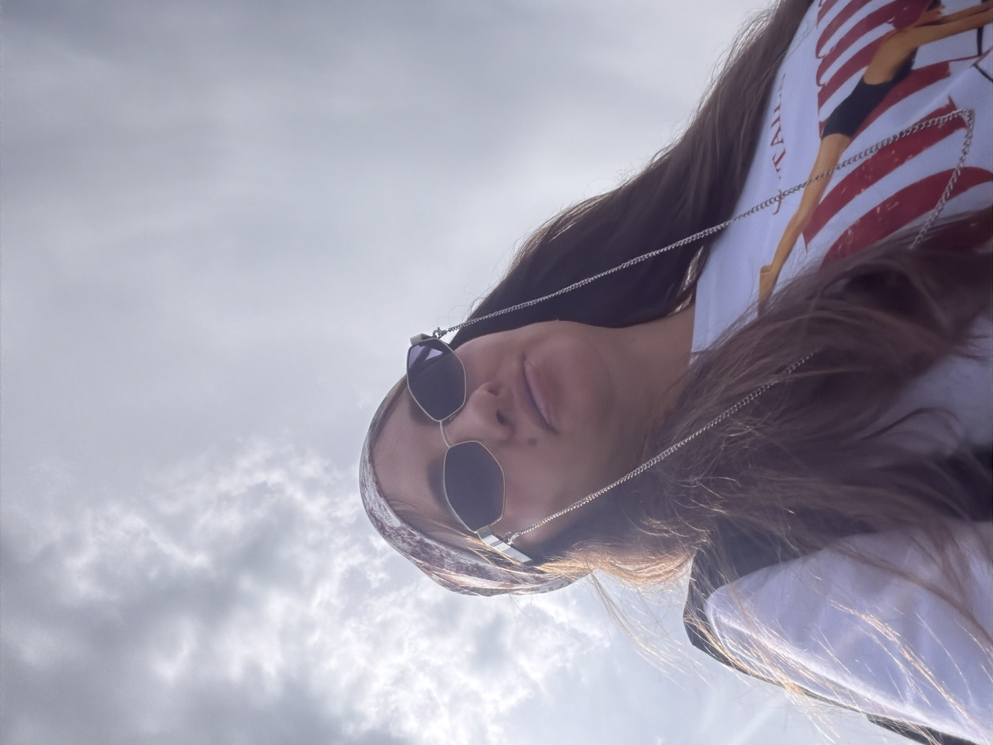 A low-angle, medium shot captures a young woman wearing sunglasses, a bandana, and a white t-shirt with a graphic design. She is looking up towards a cloudy sky. The woman's hair is brown and long, and she is wearing a silver chain around her neck that attaches to her sunglasses. Her t-shirt features a graphic of a gymnast performing a move against a backdrop of red and white stripes. The background is dominated by a cloudy sky, suggesting an outdoor setting possibly in Kremenkul', Russia, though no specific landmarks are visible. The lighting indicates it may be daytime.