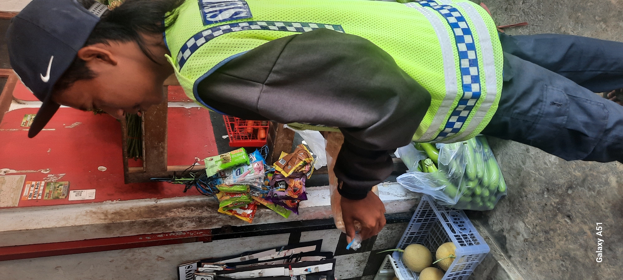 The image depicts a young man wearing a dark blue baseball cap with a white Nike logo, and a bright lime green safety vest with reflective checkered blue and white stripes over a dark gray long-sleeved shirt. He is standing on a concrete ground surface, likely in an outdoor market setting in Selopuro, Indonesia. In front of him, to his left, are two plastic crates containing light yellow cantaloupe melons. To his right, a large clear plastic bag is filled with green vegetables, possibly yardlong beans. Behind the vegetables, a small red plastic crate is visible, containing two round orange fruits. Hanging from a string is a cluster of small, individually wrapped snack packets. The background features a red wall with some peeling paint and posters, and a wooden shelf displaying more green vegetables. The man's hands are near a plastic bag, suggesting he is either interacting with or holding something within it. The overall scene suggests a casual commerce environment.