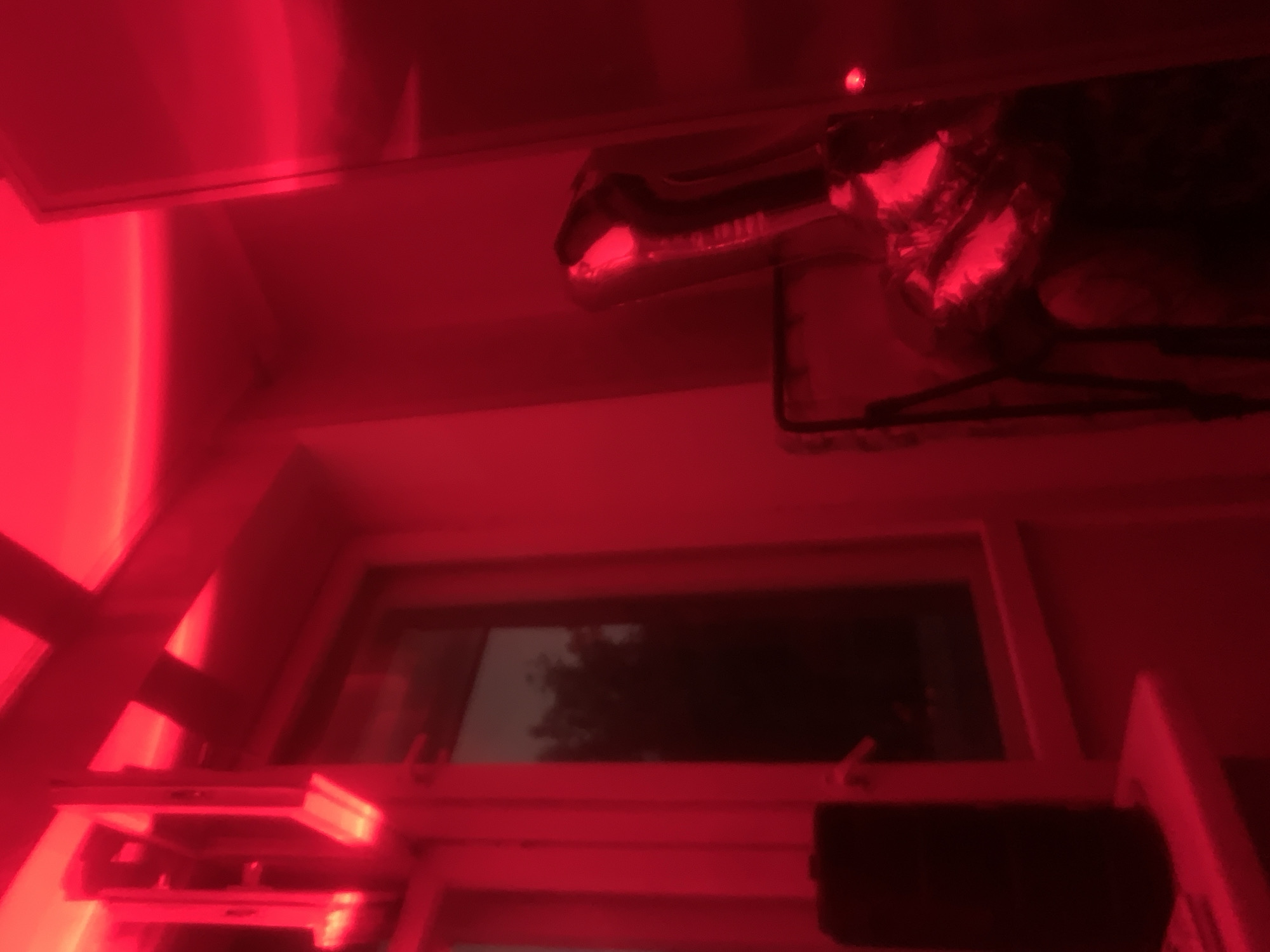 The image displays an indoor scene dominated by a strong, artificial red light. The main subject is a large, metallic red foil balloon shaped like a dinosaur or dragon head, featuring visible teeth and an open mouth. This balloon appears to be suspended in the upper right portion of the frame, possibly from a hanger or support structure which is also visible. A small, bright red point of light is discernible above the balloon.
Below the balloon, a window offers a view to the outside, which is mostly dark but shows vague silhouettes that suggest trees against a dim sky. The strong red lighting originates from within the room, making it difficult to discern the actual time of day outdoors, though the dimness through the window implies it might be evening or night. The room itself appears to be an ordinary interior space, with visible walls, a ceiling, and a window frame. In the lower right corner, a dark, rectangular object, possibly an electronic device, rests on a surface.
No activity or event is taking place; the scene is static. The intense red illumination creates a distinct, moody, or thematic atmosphere within the room. There is no visible text in the image.
