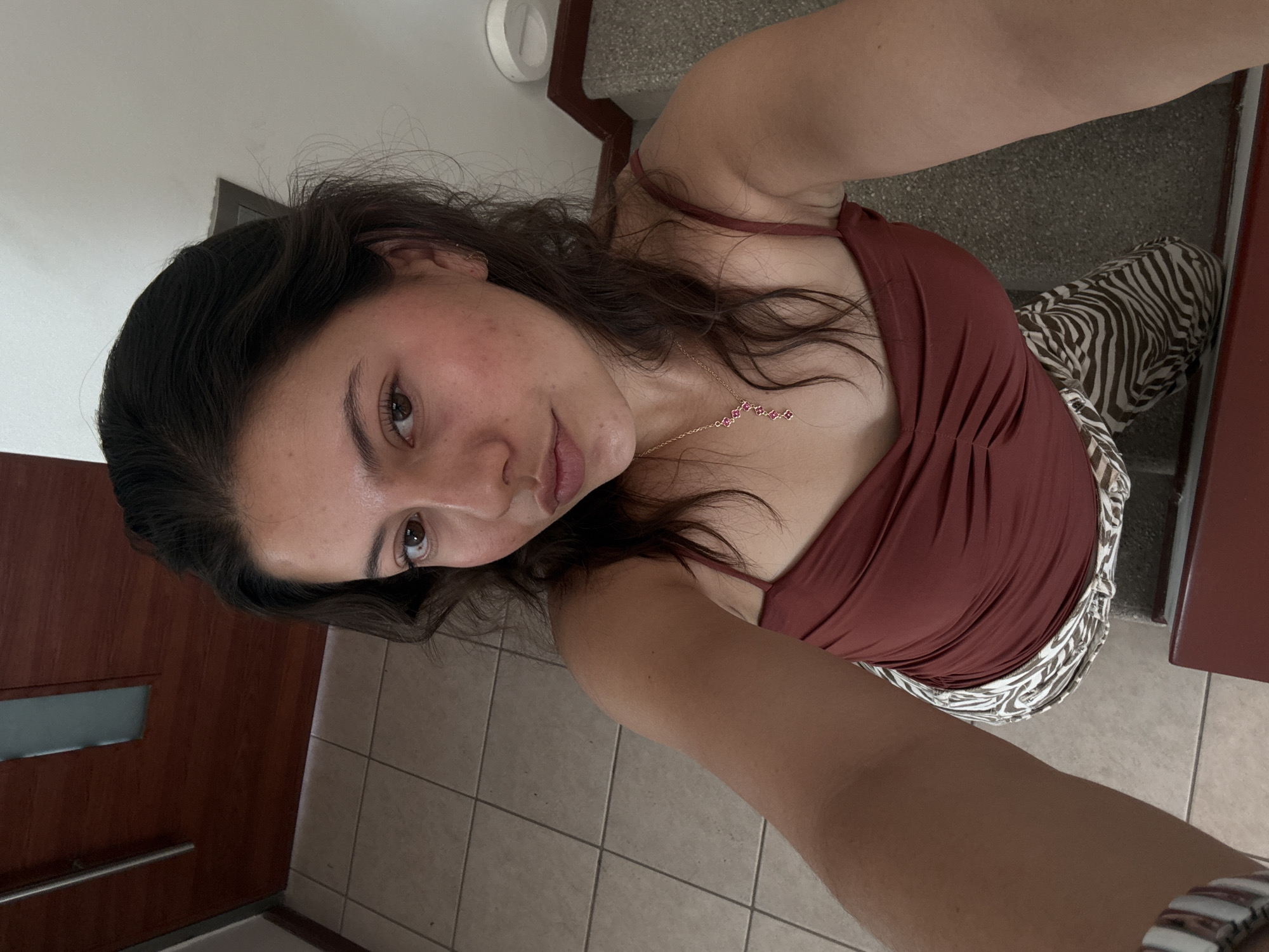 A young woman is taking a selfie indoors. She is wearing a maroon-colored tank top and zebra-striped pants. She is also wearing a delicate gold necklace with small pink flower-like details. She has brown hair and light brown eyes. She appears to be standing near a set of stairs. The floor is tiled, and a portion of a wooden door is visible in the background. The exact location in Heroica Puebla de Zaragoza, Mexico cannot be definitively determined from the provided image.