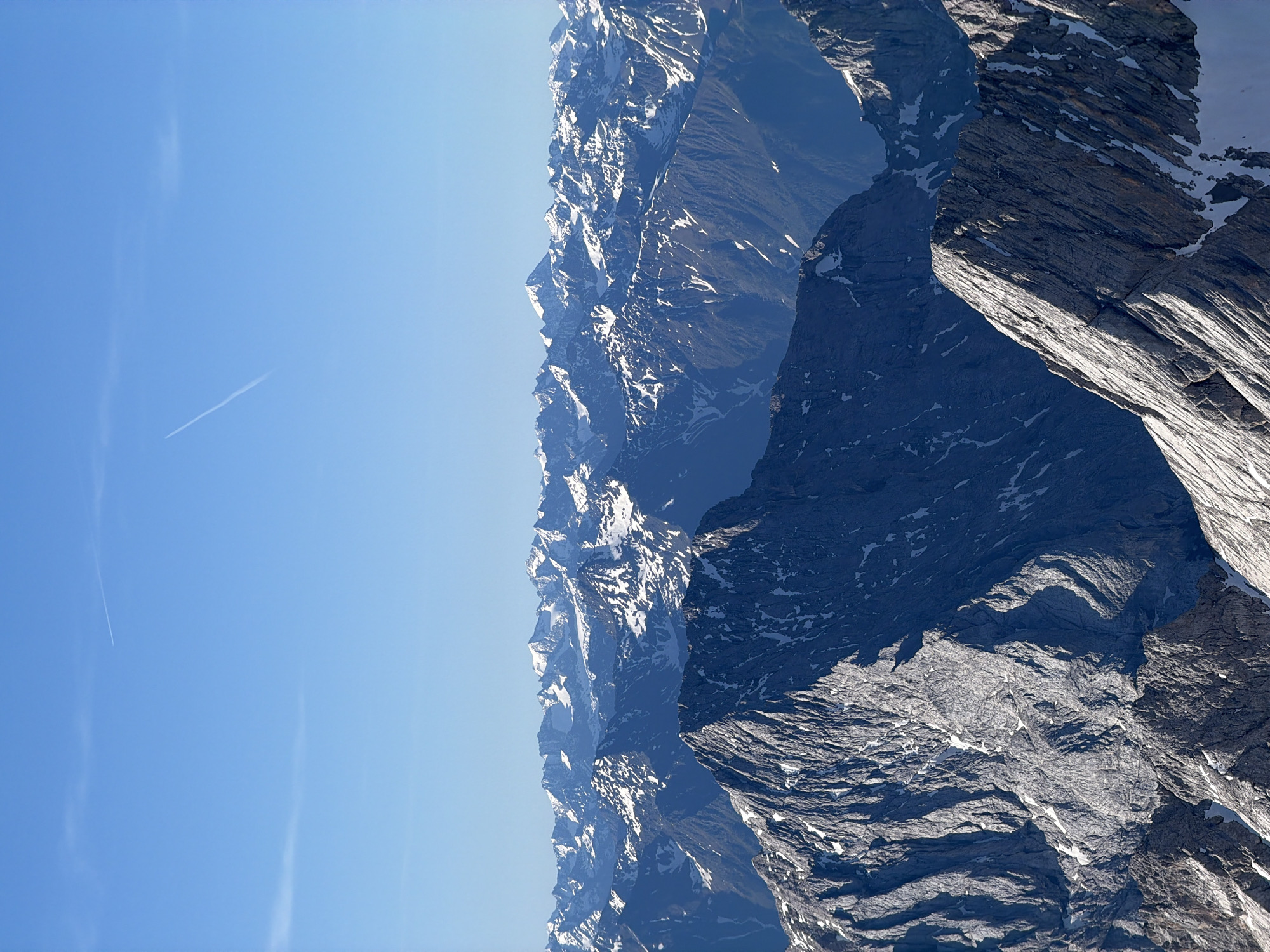 The image depicts a panoramic view of snow-capped mountains under a clear blue sky. Two faint contrails from airplanes are visible in the upper sky. In the foreground, the jagged, rocky peaks of mountains are prominent, with patches of snow clinging to their surfaces. The midground and background reveal a vast expanse of layered mountain ranges, also dusted with snow. The sunlight illuminates the scene, casting shadows that accentuate the rugged topography. This is a high-altitude, mountainous landscape, likely taken from a vantage point offering an extensive view of the Alps. Given the description of Grainau, Germany, this scene is likely of the Bavarian Alps, possibly featuring the Zugspitze massif, Germany's highest peak, which is near Grainau. There are no people or man-made structures visible in the image, focusing entirely on the natural environment.