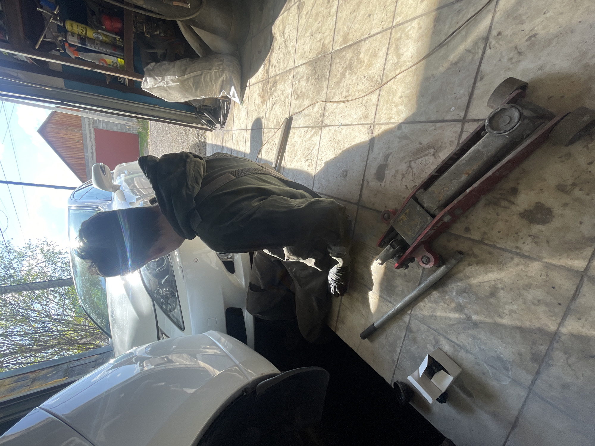 A person wearing dark green coveralls kneels next to a white car inside a garage in Минино, Russia. The person is facing the front of the car, appearing to be working on the vehicle. Next to the person, on the concrete floor of the garage, is a red hydraulic floor jack with its handle adjacent to it. There is also a small, white, box-like object on the floor nearby. The garage door is partially open, providing sunlight into the scene. Items are stored on shelves near the top of the garage opening.