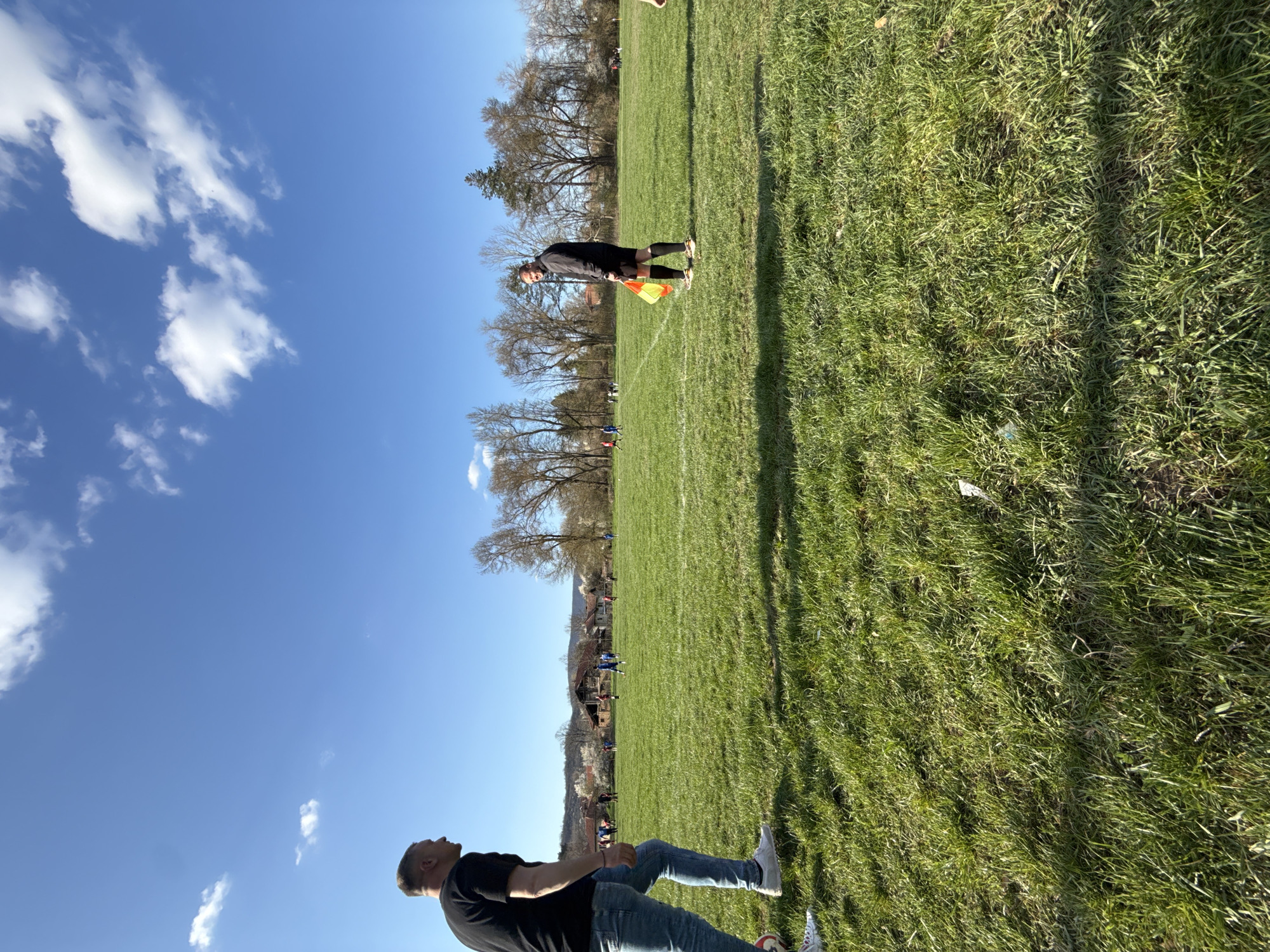 A sunny outdoor scene captures a soccer match in Dushevo, Bulgaria. The blue sky is dotted with scattered clouds. Two men are visible on the sidelines: one is standing with a flag in hand, clad in a black uniform, while the other is seated, wearing a black shirt and blue jeans. The soccer field is well-maintained with green grass and white lines. In the background, trees and houses are visible, suggesting a rural or suburban environment. The players are visible on the field.