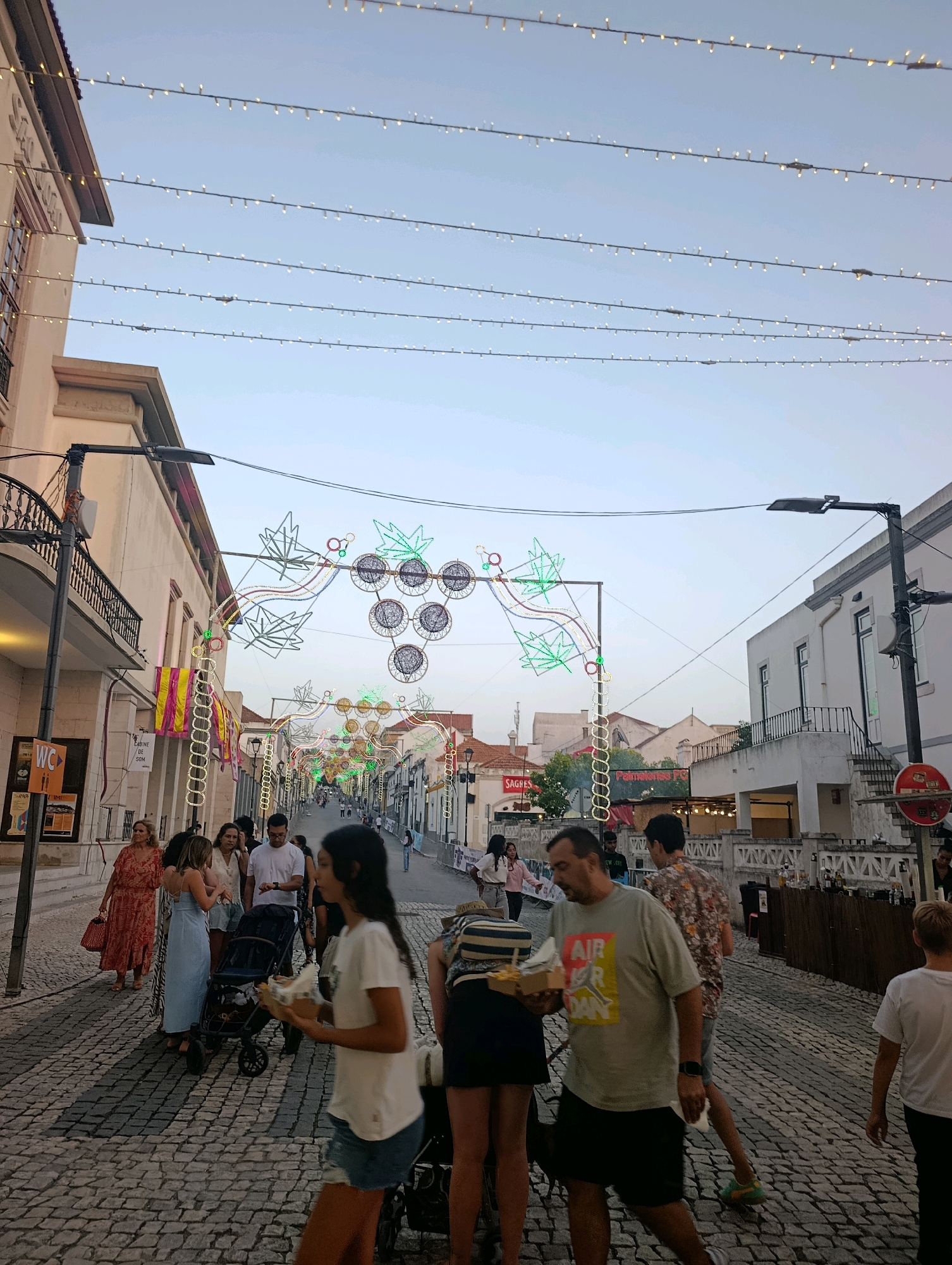 The image depicts a street scene in Palmela, Portugal, at dusk. The street is paved with cobblestones and lined with buildings on both sides. String lights are strung across the street, illuminating the scene and creating a festive atmosphere. In the foreground, several people are walking, some with food in hand. In the background, a large, illuminated archway with decorative grape clusters is visible, suggesting a festival or celebration is taking place. The buildings appear to be old and traditional, with balconies and shutters. The sky is a pale blue, indicating the end of daylight.