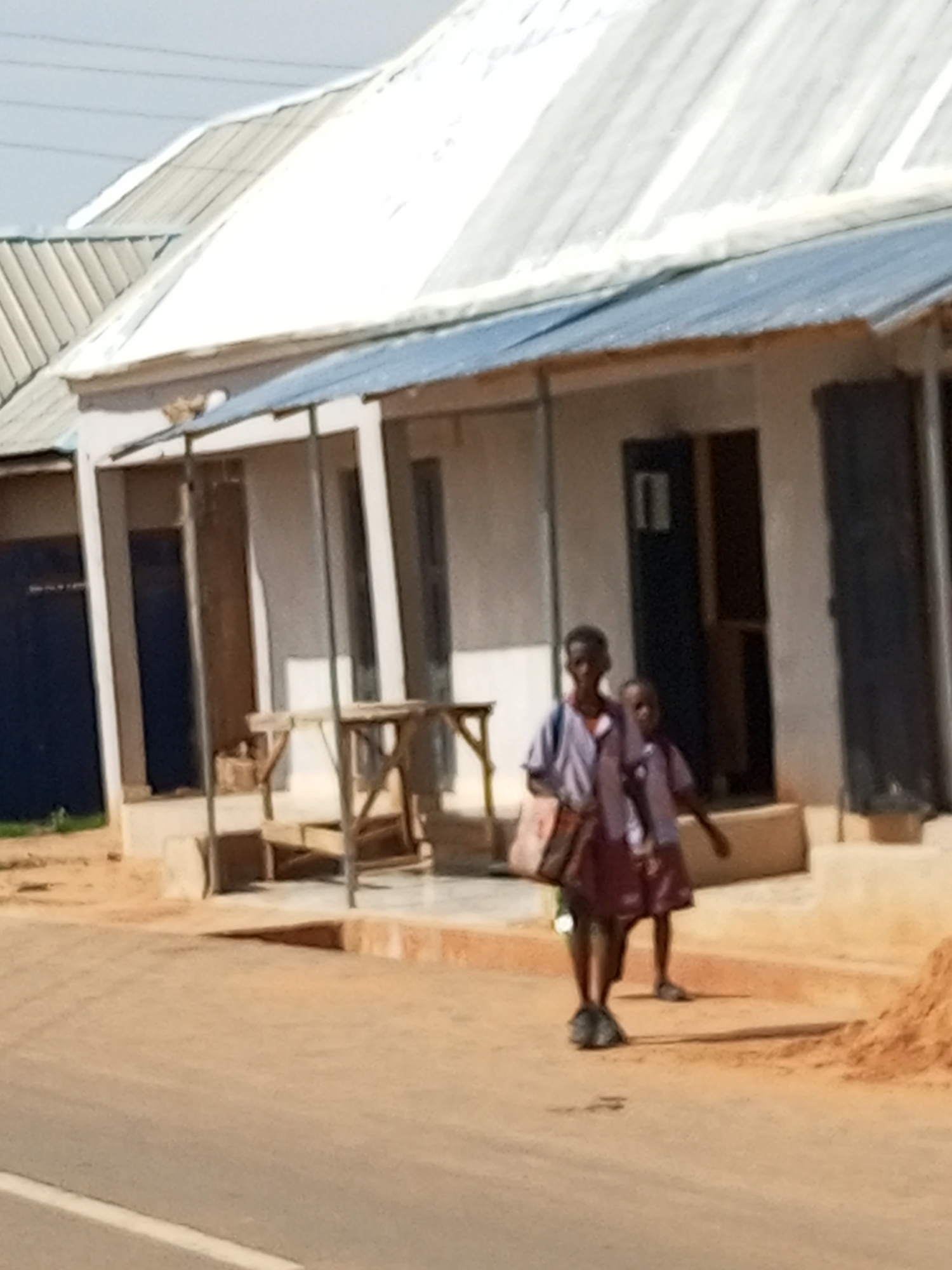 Two young students, one taller than the other, walk together on a dirt road in front of a row of buildings. Both are wearing uniforms consisting of a white and purple striped shirt with a dark skirt for the taller student and a dark skirt for the shorter student. The taller student carries a patterned bag over their left shoulder. The buildings have light-colored walls and corrugated metal roofs, one with a blue trim. A wooden stall or table is set up in front of one of the buildings. The ground is a mixture of dirt and sand, with some scattered debris. The sky is clear and bright. The scene appears to be in a town or city, possibly Gombe, Nigeria, given the architectural style and environment.