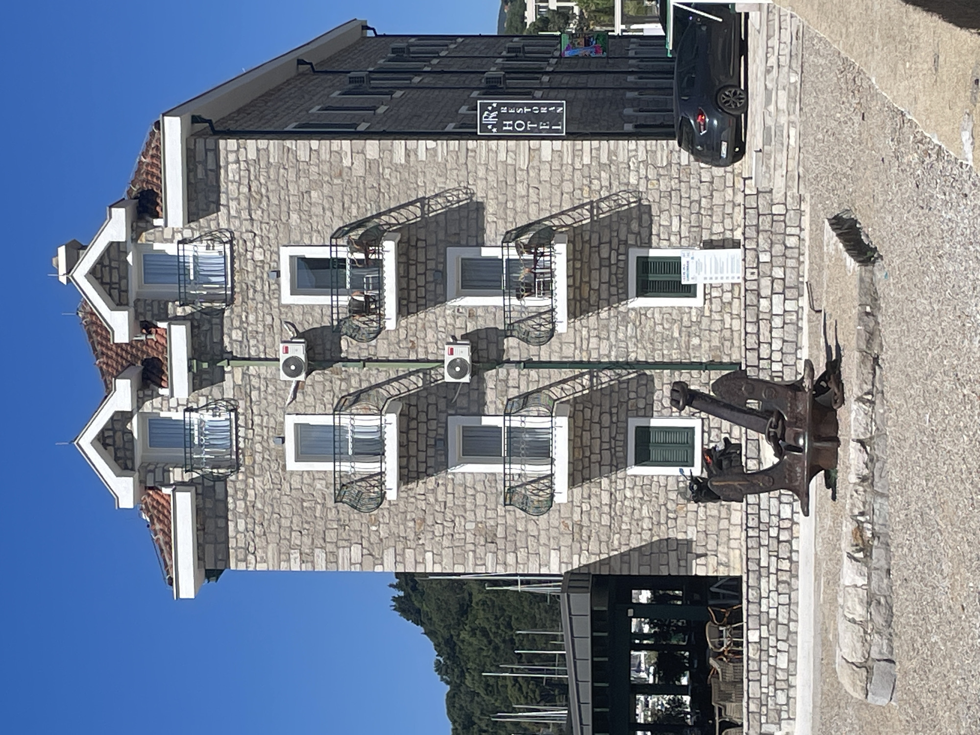 The image depicts a stone-clad hotel in Meljine, Montenegro. The hotel is several stories tall, featuring gabled roof sections, white-framed windows with decorative metal balconies, and green-shuttered windows. Air conditioning units are mounted on the exterior walls. Above the top windows, a sign identifies the building as a hotel. A dark-colored car is parked near the hotel's entrance, which is paved with stone. A large, old anchor sits on the pavement near an outdoor dining area, which suggests a nautical theme. Vegetation and the side of a building are visible in the background. The sky is clear and blue.