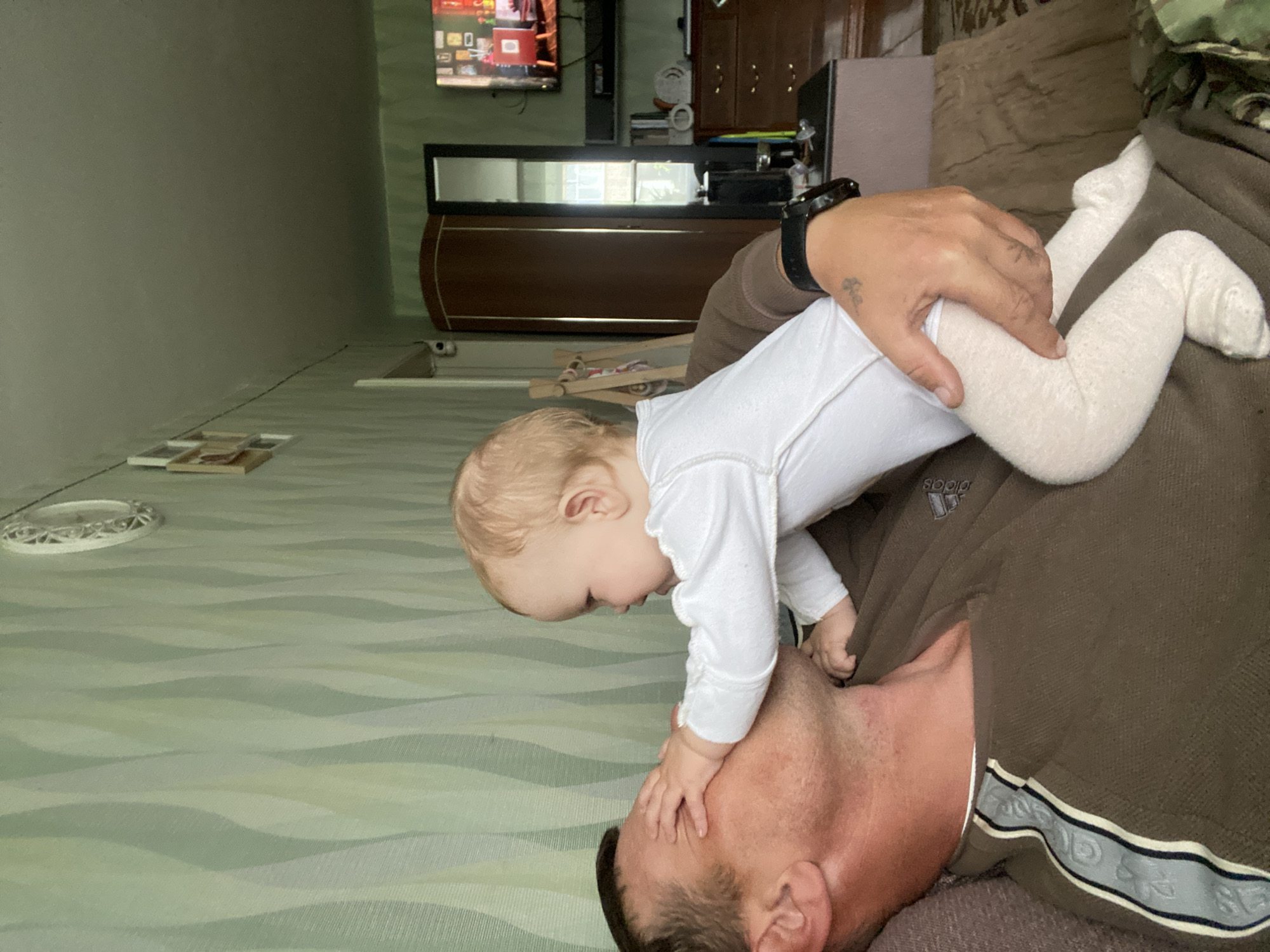 An adult male and an infant are present in an indoor residential setting in Фоки, Russia. The male, with short brown hair, lies on his back on a brown upholstered surface. He wears a dark brown long-sleeved shirt featuring a light blue and white stripe with an "adidas" logo on the left sleeve, and a black wristwatch on his left wrist. An infant, with light blonde hair, is positioned on top of the man, facing him. The infant wears a white long-sleeved bodysuit and light-colored tights. The infant's right hand covers the man's upper face, while the man's left hand supports the infant's left leg.
The room features light green wallpaper with a vertical wavy pattern. The ceiling is a light grey-green. On the upper left wall, two small, framed items are visible, connected by a thin black cable that runs along the wall to the right. On the upper right, a white ornate wall clock is mounted. A white dome-shaped security camera is affixed high on the wall in the central background, above a tall, dark brown wooden wardrobe with gold trim. Adjacent to the wardrobe is a tall, black-framed mirror reflecting an exterior window with vertical elements. To the right of the mirror, a flat-screen television displays a colorful image on a wall-mounted unit. Below the television is a dark brown wooden dresser. Various items, including a white clock, are on the dresser and a dark surface in front of it, where a pacifier and other small objects are visible. A camouflage-patterned fabric is partially visible on the lower right of the frame.