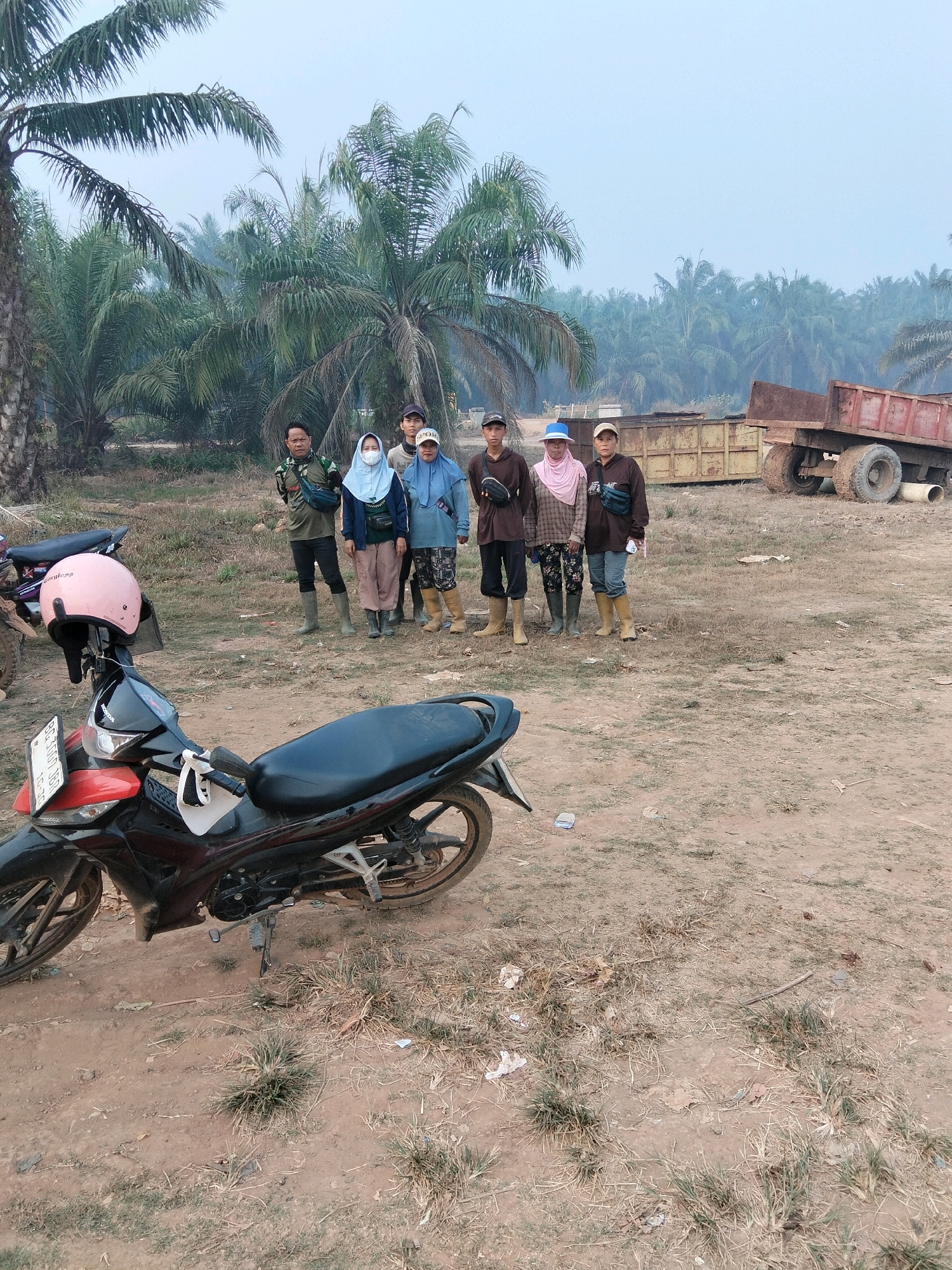 In Teluk Tenggirik, Indonesia, a group of seven people stands in an outdoor area with palm trees in the background and a red truck in the distance. A motorcycle is parked in the foreground. The group is composed of four men and three women. The men are wearing casual clothing, including t-shirts, long pants, and hats. Two of the women are wearing headscarves, and one is wearing a face mask. They are all wearing boots. The environment is a dirt and grass area, likely a plantation or rural location. The sky is overcast and hazy, suggesting a dry or dusty atmosphere.