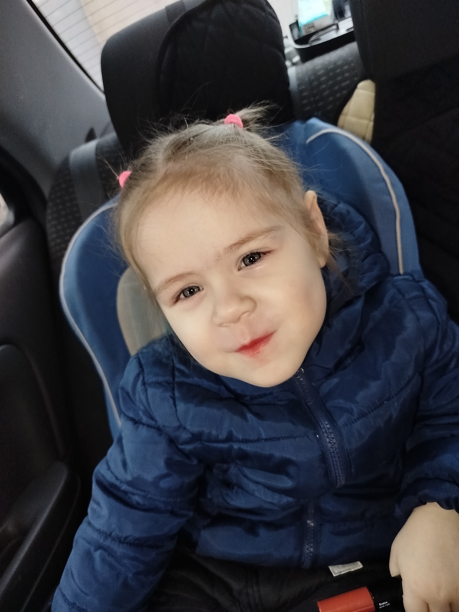 A young child, approximately a toddler, is seated in a blue car seat within a vehicle in Chaikovsky, Russia. The child has light-colored hair, styled with two pink hair ties visible on the top of the head, and dark eyes. A slight, closed-lip smile is present, with a reddish tint on the lips. The child is wearing a dark blue, possibly padded or quilted, jacket with a front zipper and visible hood. Dark fabric, likely trousers, is visible below the jacket. The car's interior, featuring dark-colored seats and headrests, forms the background. A blurred light source is visible through the rear window. A red object with the Russian text "Блеск для губ" (Lip Gloss) is partially visible in the bottom right corner of the frame, near the child's lap.