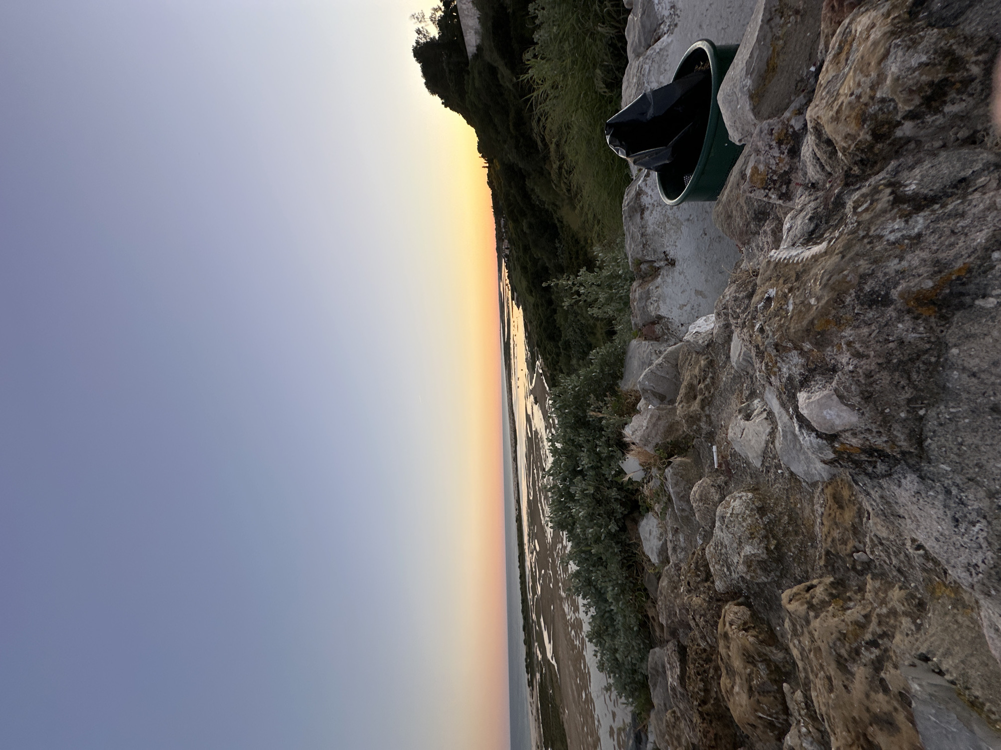 The image shows a landscape at the mouth of the Guadiana River in Vila Real de Santo António, Portugal, during dusk or dawn. The sky transitions from blue to orange and yellow near the horizon. The river, partially exposed with sandbanks, is visible in the middle ground. In the foreground, a rough stone wall is topped with vegetation and a green trash can containing a black bag.