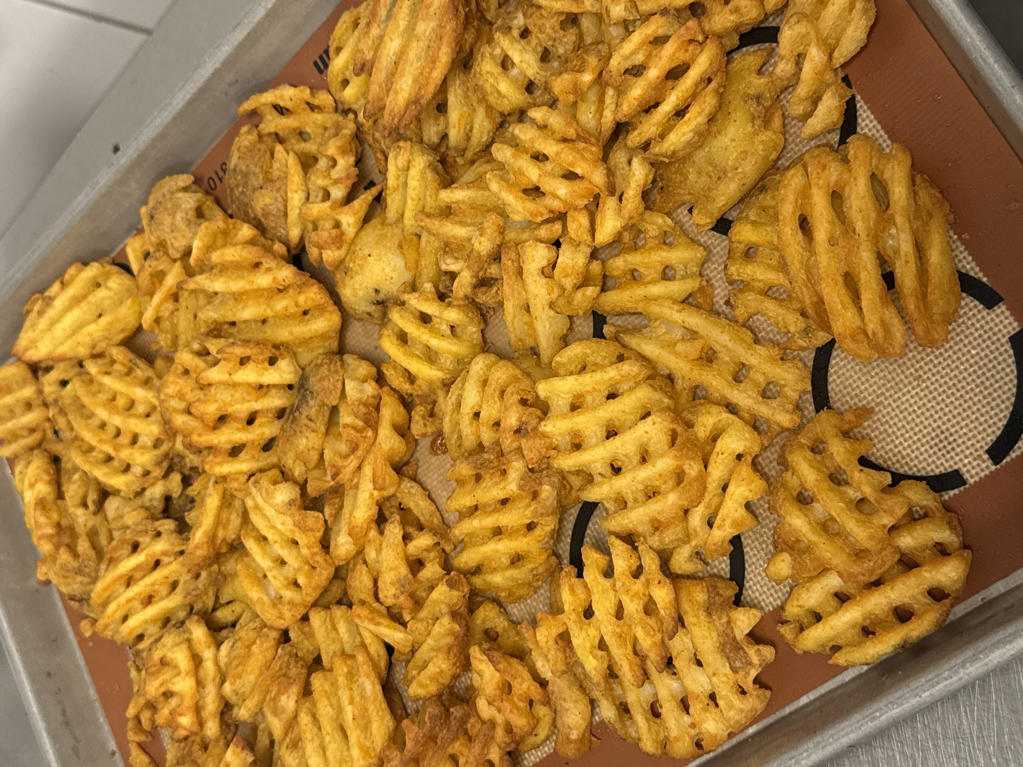 A close-up overhead shot shows a baking tray filled with waffle-cut french fries. The fries are golden brown with some darker, crispy edges and appear to be seasoned with paprika or a similar reddish spice. They are spread across a brown silicone baking mat, which is placed on top of a metallic baking tray. The background is blurred, suggesting an indoor setting, possibly a kitchen. No people or specific city details are visible.