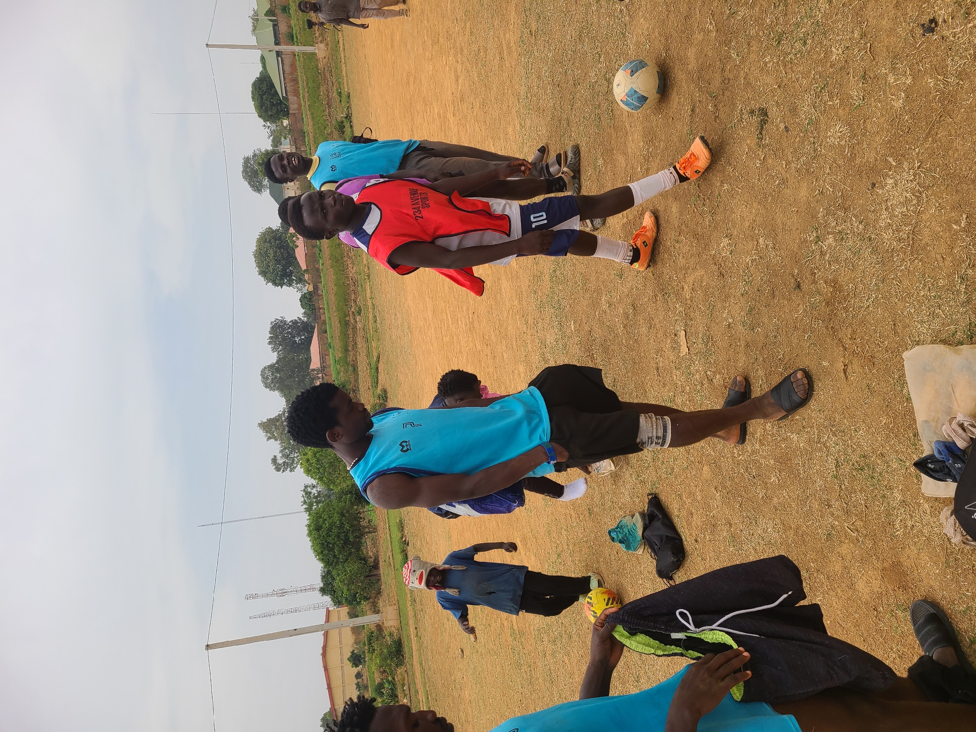 The image shows a group of people on a dirt field in Barikin Jirga Summa, Nigeria. The field has sparse vegetation. Three men are standing in a line near a soccer ball. One man is wearing a red vest with the number 10. Another man is wearing a blue sleeveless shirt. Other individuals are present on the field. Clothing items are scattered on the ground. Tall metal structures are visible in the distance.