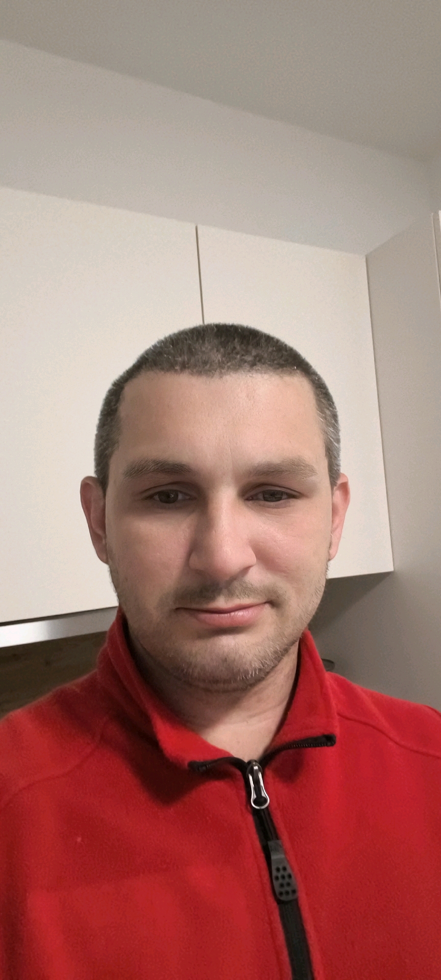 The image shows a close-up, frontal view of a man with short, dark hair and light skin. He has brown eyes and a slight stubble. He is wearing a bright red zippered jacket. The background appears to be a domestic setting with light-colored cabinets above him. The lighting is neutral, and there is no visible indication of his specific location within a city.