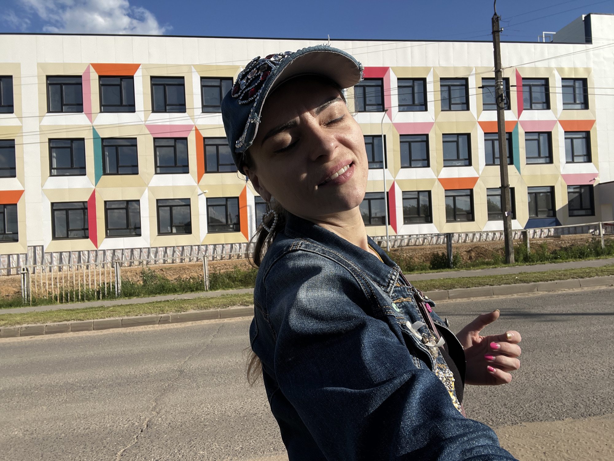 A woman stands outside in front of a modern building with a colorful facade. The building has a grid pattern of windows framed by geometric shapes in shades of yellow, orange, pink, and teal. The woman is wearing a denim jacket and a denim baseball cap adorned with rhinestones and beads. Her eyes are closed, and she has a slight smile, suggesting she is enjoying the moment or the sunshine. She is wearing heart-shaped earrings and has bright pink nail polish on her fingers, with one finger having gold nail polish. Her right hand is slightly raised, with her thumb extended, as if hitchhiking or signaling. The scene appears to be outdoors during the day, with a clear blue sky and some clouds visible. The ground in front of the building is a sidewalk and a road, with a grassy area and a simple metal fence separating the sidewalk from the building. A utility pole stands near the building. The overall impression is one of casual relaxation and perhaps a sense of travel or waiting. The location is identified as Krasnoe-na-Volge, Russia.