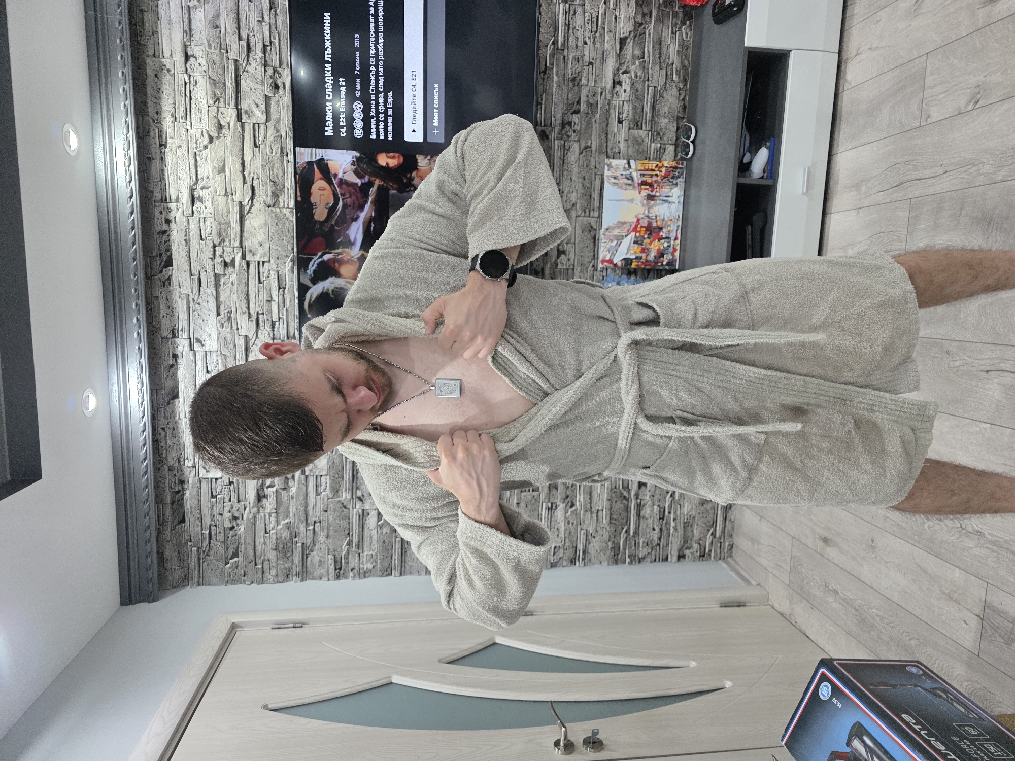 A man in a robe stands in a room. He is adjusting his robe and wearing a watch. The room has a brick wall, a television playing a Bulgarian TV show, a cabinet, a door, and wood flooring. A box is on the floor in the foreground. The image does not contain enough information to determine the location in the city of Yagodovo, Bulgaria, if it is actually there.