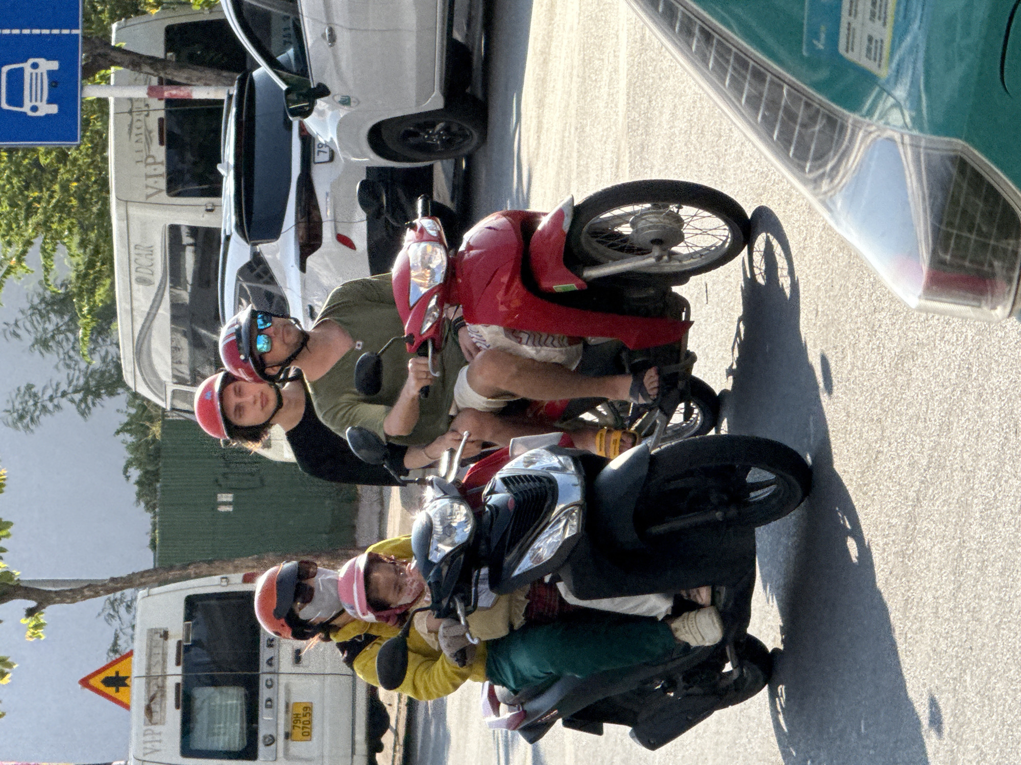 The image shows a group of people riding on motorcycles in what appears to be a street scene in Nam Nha Trang Ward, Vietnam. The primary subjects are the individuals on the motorcycles and the vehicles themselves. There are at least three motorcycles visible. The first, a red scooter, carries two people wearing helmets. The rider is a person wearing a green shirt and blue sunglasses, and behind them is another person wearing a red helmet and black top. The second motorcycle, a darker color, also has multiple riders. The person in front is wearing a pink helmet and a yellow jacket. Another person behind them is wearing a pink helmet, a grey scarf, and a yellow jacket. The scene is outdoors, on an asphalt road. The lighting suggests it is daytime, possibly late morning or early afternoon, with strong sunlight casting shadows. In the background, there is a white van with "DCAR" visible on the side and a license plate that reads "79H 070.59". A yellow triangular road sign with a black cross is also visible. To the left of the scene, a blue sign with a white drawing of a car and dashed lines is present. The overall atmosphere seems to be a busy street with vehicles and pedestrians. The individuals appear focused on navigating the road, suggesting everyday commuting or travel. The weather appears to be clear and sunny.