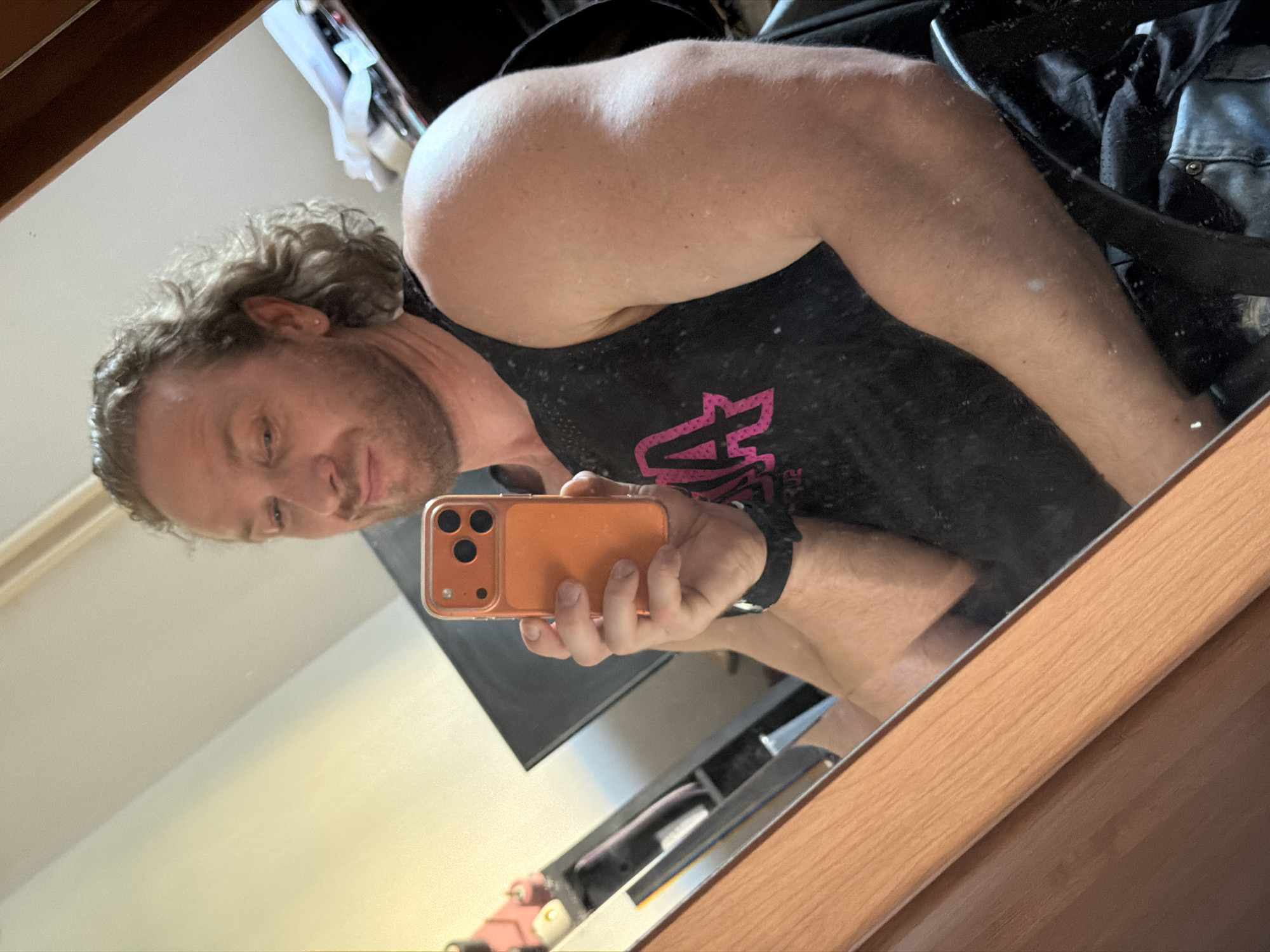 A man is depicted taking a mirror selfie indoors. He has medium-length curly blonde hair and a short beard, wearing a black sleeveless tank top with a partial pink graphic resembling "ZUMBA BA 02". He holds an orange-cased smartphone with a triple camera array in his left hand, capturing his reflection. His expression is a gentle smirk, and his right arm is slightly flexed. A small stud earring is visible in his left ear, and he wears a black watch on his left wrist. The mirrored background reveals a room with light-colored walls, a dark piece of furniture, and various objects including papers and a pink item on shelves. A wooden frame borders the bottom left edge of the mirror. There is no visual information within this image to confirm its location in the city of Saint Petersburg, Russia.