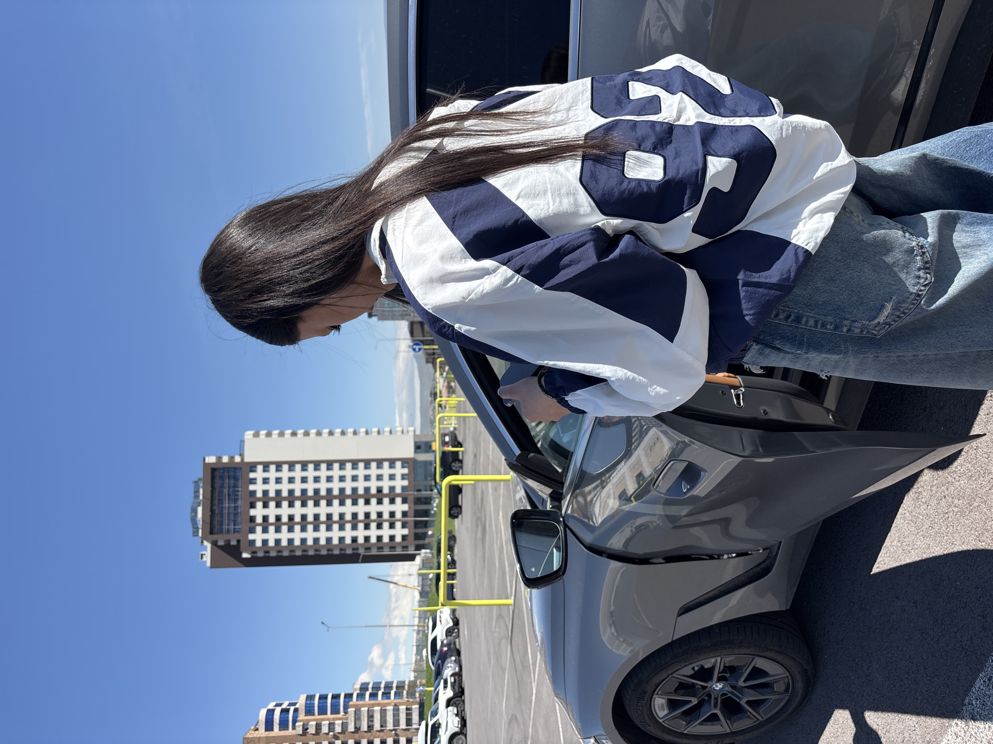 A young woman with long, dark hair is standing next to a gray car with the door open, possibly about to get in or out. She is wearing a white and blue jacket with the number "88" on the back and blue jeans. The car is parked in a parking lot with yellow bollards. In the background, there are two buildings, one taller than the other, suggesting an urban setting. The sky is clear and blue, indicating a sunny day. Based on the architectural style of the buildings, the location is identified as Yerevan, Armenia.