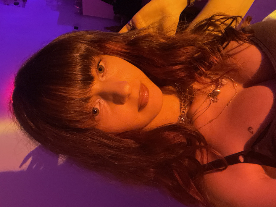 A woman with auburn hair and bangs is lying down. Her face is turned to the camera, with eyes open and lips slightly parted. She is wearing a black top with thin straps and several necklaces. The lighting is a mix of purple and orange hues, creating a warm and vibrant atmosphere. Her skin appears smooth and her expression is relaxed.