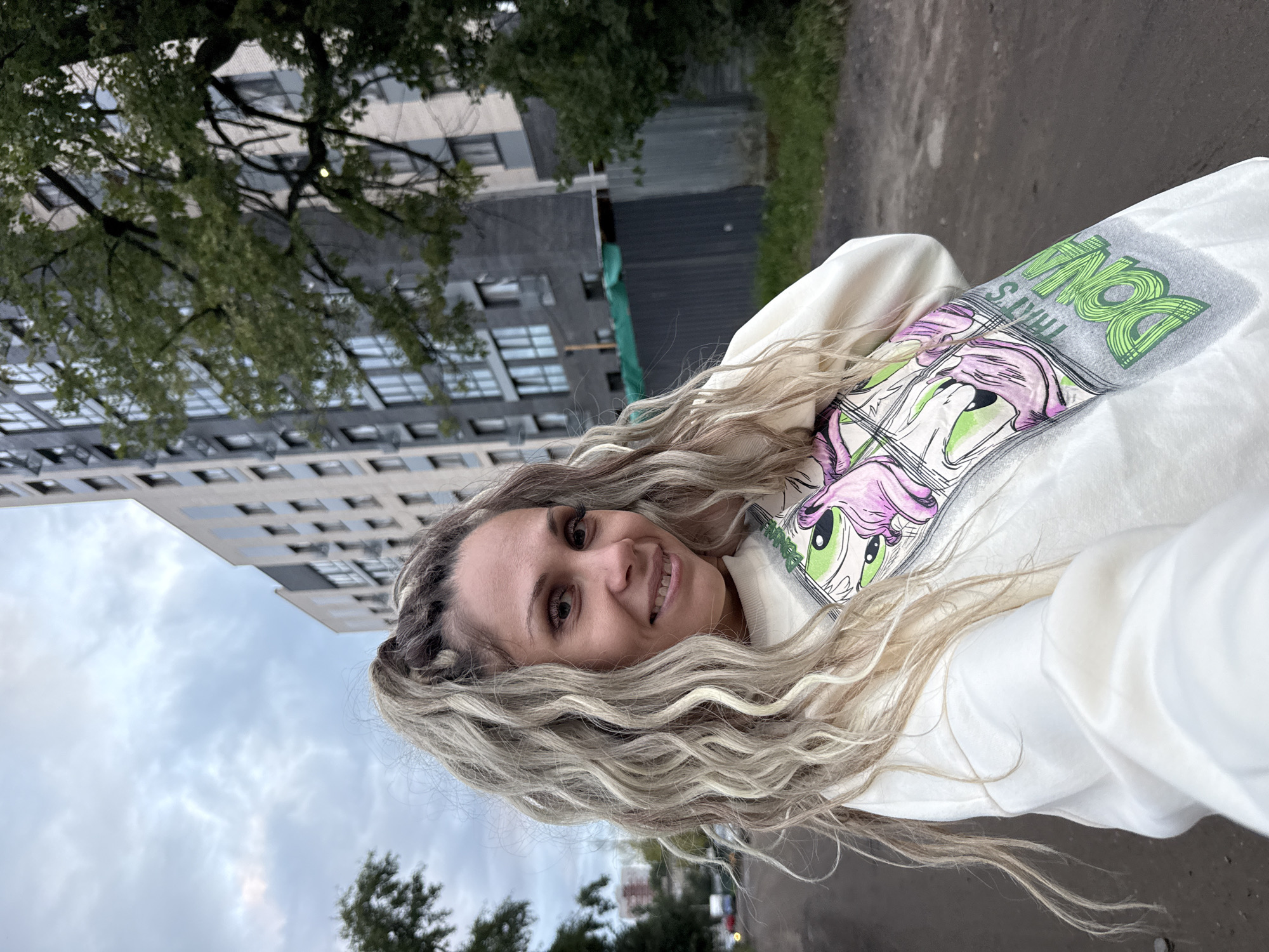 A woman with long, wavy blonde hair pulled into a high ponytail stands in an outdoor urban setting. She is wearing a white sweatshirt with a graphic print on the front. The background features a tall, modern apartment building and a mature tree with green foliage. The ground is a dirt path. The sky is overcast. This scene is located in Rusanovo, Russia.