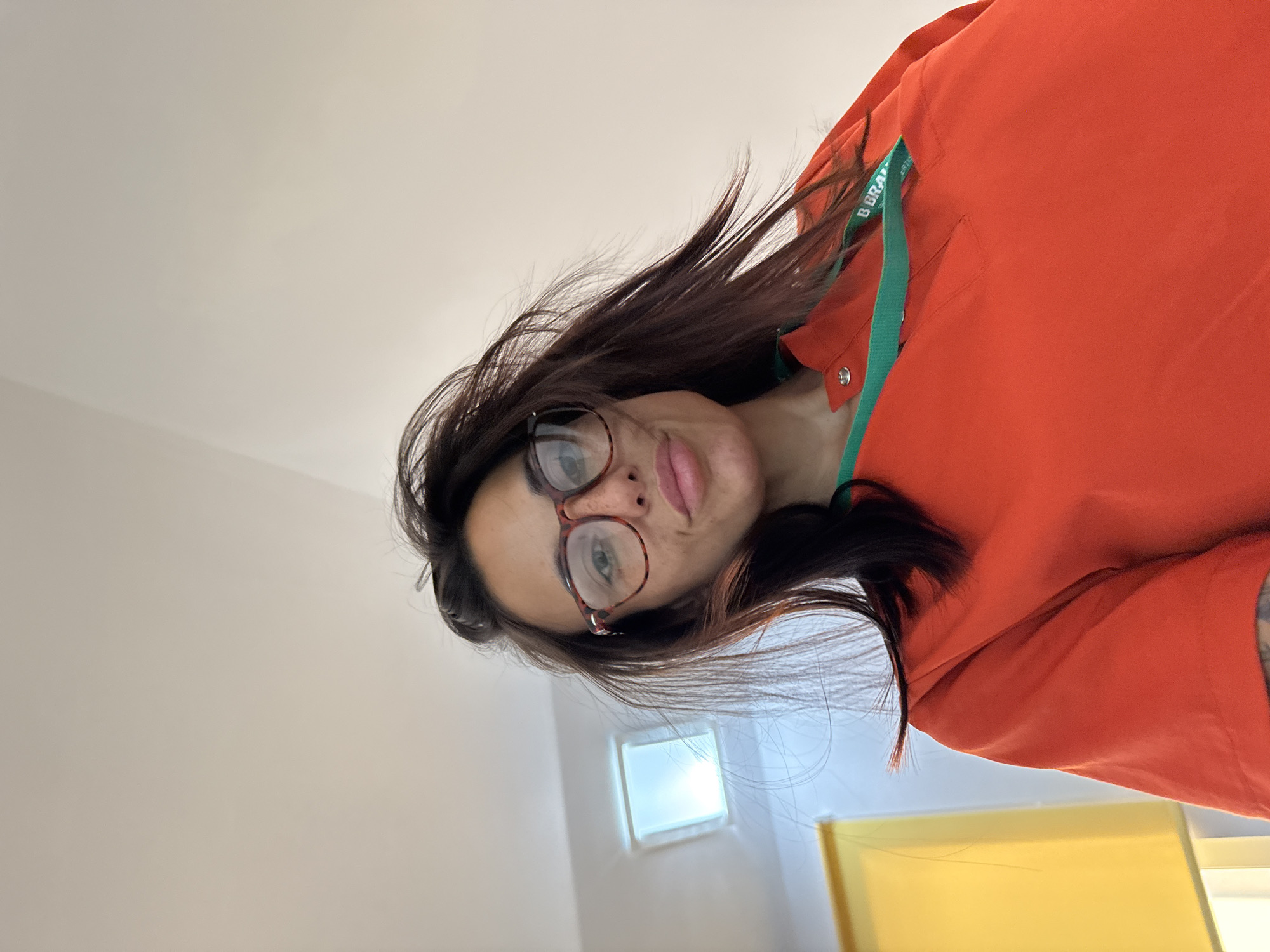 A woman with long, dark hair and glasses is taking a selfie from a low angle. She is wearing a bright orange shirt with a green trim around the collar and a lanyard. Her hair is windswept. In the background, there is a skylight and a yellow blind. The scene is indoors.
