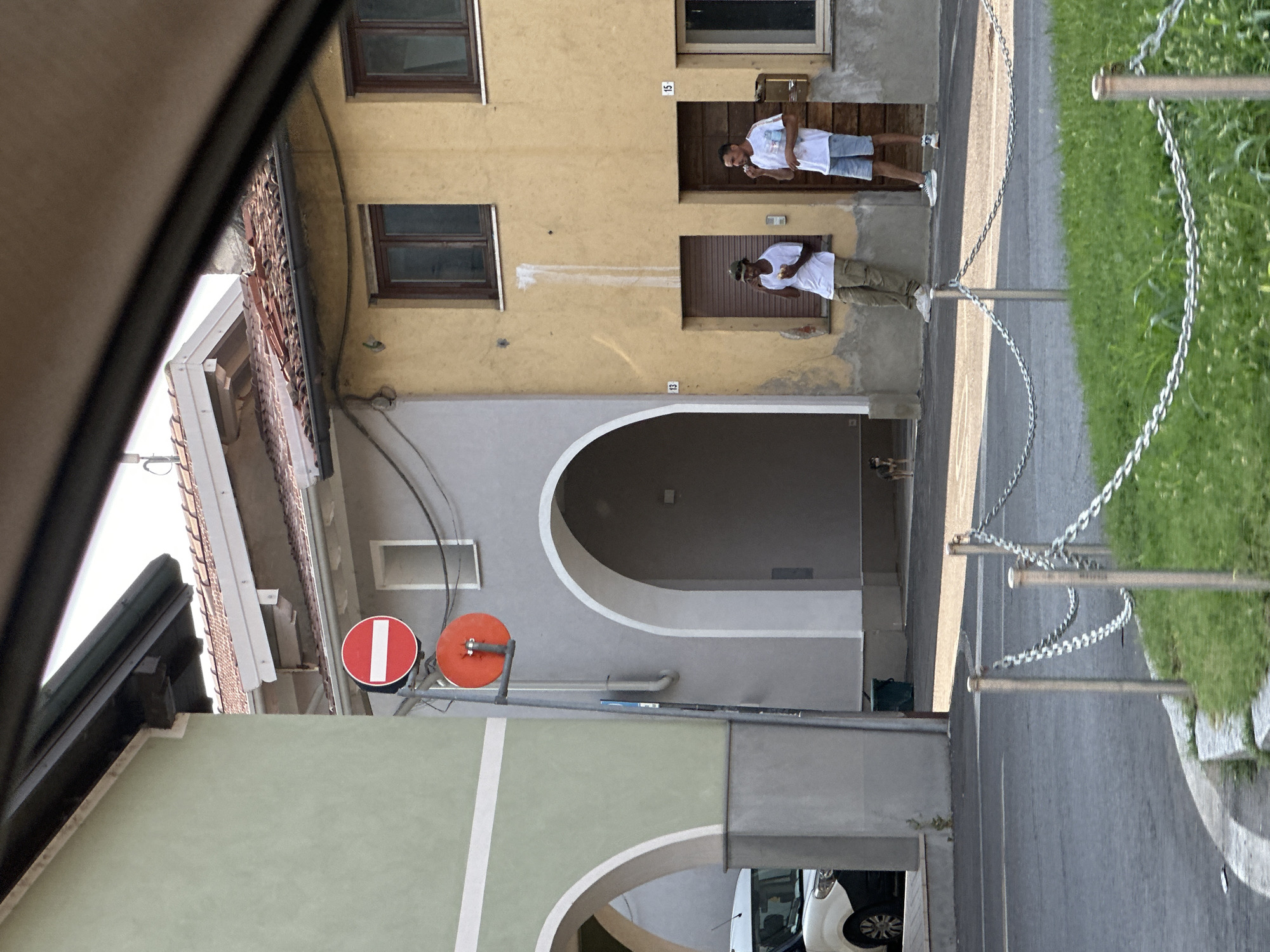 Two individuals are captured in a medium shot, standing on a street outside a building. The individual on the left, dressed in a white t-shirt and blue shorts, is holding a white object to their mouth. The person on the right wears a white t-shirt and khaki pants, also with a white object near their mouth. They appear to be in an urban or semi-urban setting, possibly in Rudiano, Italy, indicated by the architectural style of the buildings and the presence of chain barriers alongside a grassy area and a paved road. The time of day appears to be daytime, with bright sunlight casting shadows. One building features visible house numbers "15" and "13". Red and orange circular signs are mounted on a pole. In the background, parts of other buildings and a white vehicle are visible. The scene suggests a casual moment, with the individuals engaged in an activity that appears to involve eating or drinking.